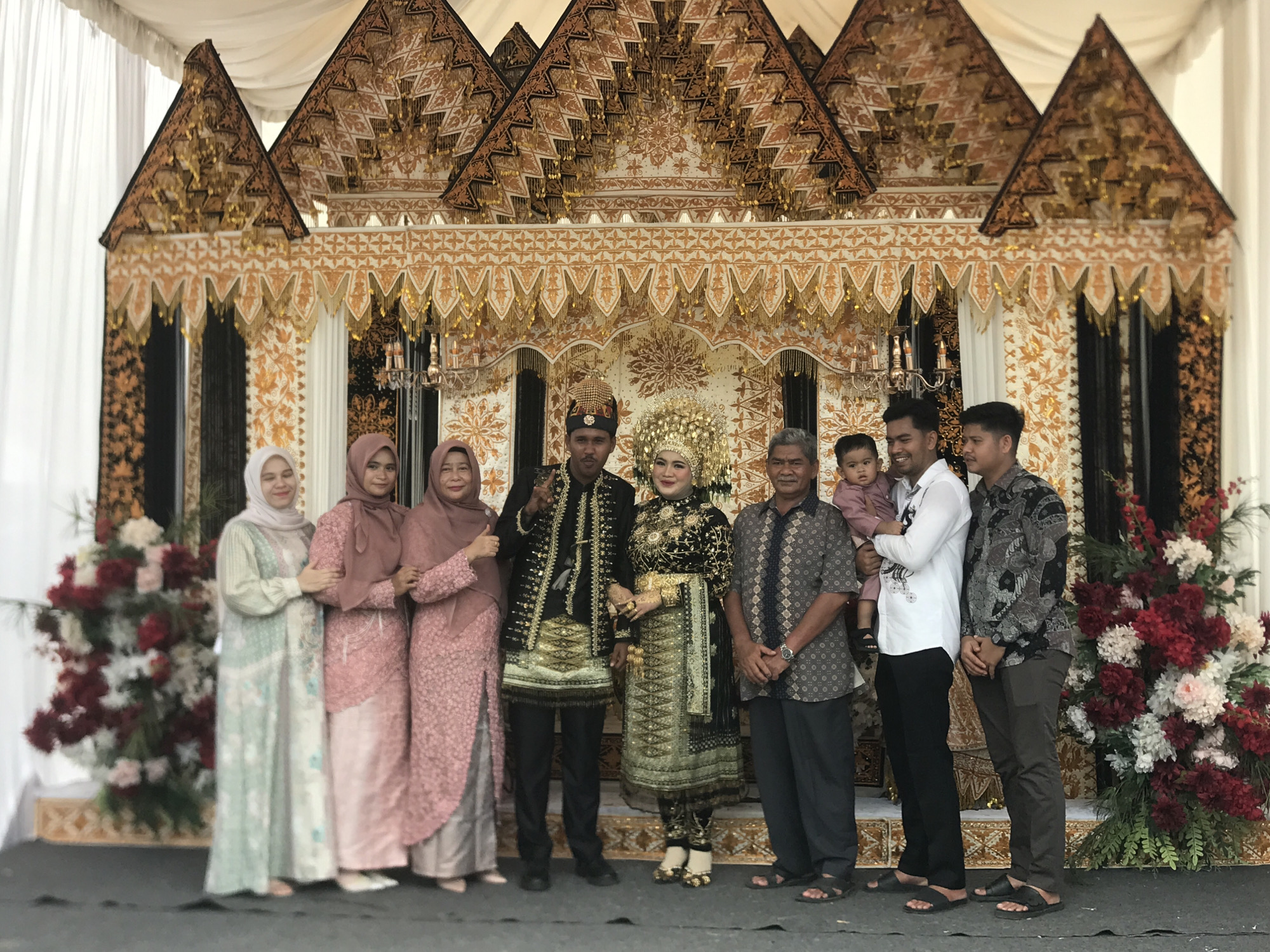 A photograph taken in Cotgapu, Indonesia, shows a group of people posing in front of a decorated backdrop. The backdrop has a decorative structure overhead resembling tiered roofs, with intricate gold and white patterns.
To the left, three women stand in a row, all wearing hijabs and long dresses. The woman on the far left wears a light green dress, the one in the middle wears a pink dress, and the woman on the right wears a light pink dress.
Next to them is a man in traditional attire, wearing a black outfit with gold accents. He has a decorative hat and is holding up a peace sign with his left hand. Standing beside him is a woman in similar traditional garb, wearing a gold headdress and a black and gold dress.
To the right of the bride is an older man wearing a patterned shirt and dark pants, with his hands clasped in front of him. Beside him is a younger man holding a baby in a pink outfit. The next man over is wearing a white shirt and black pants. Finally, on the right, another man in a patterned black and white shirt and brown pants completes the line-up.
On either side of the group are floral arrangements.