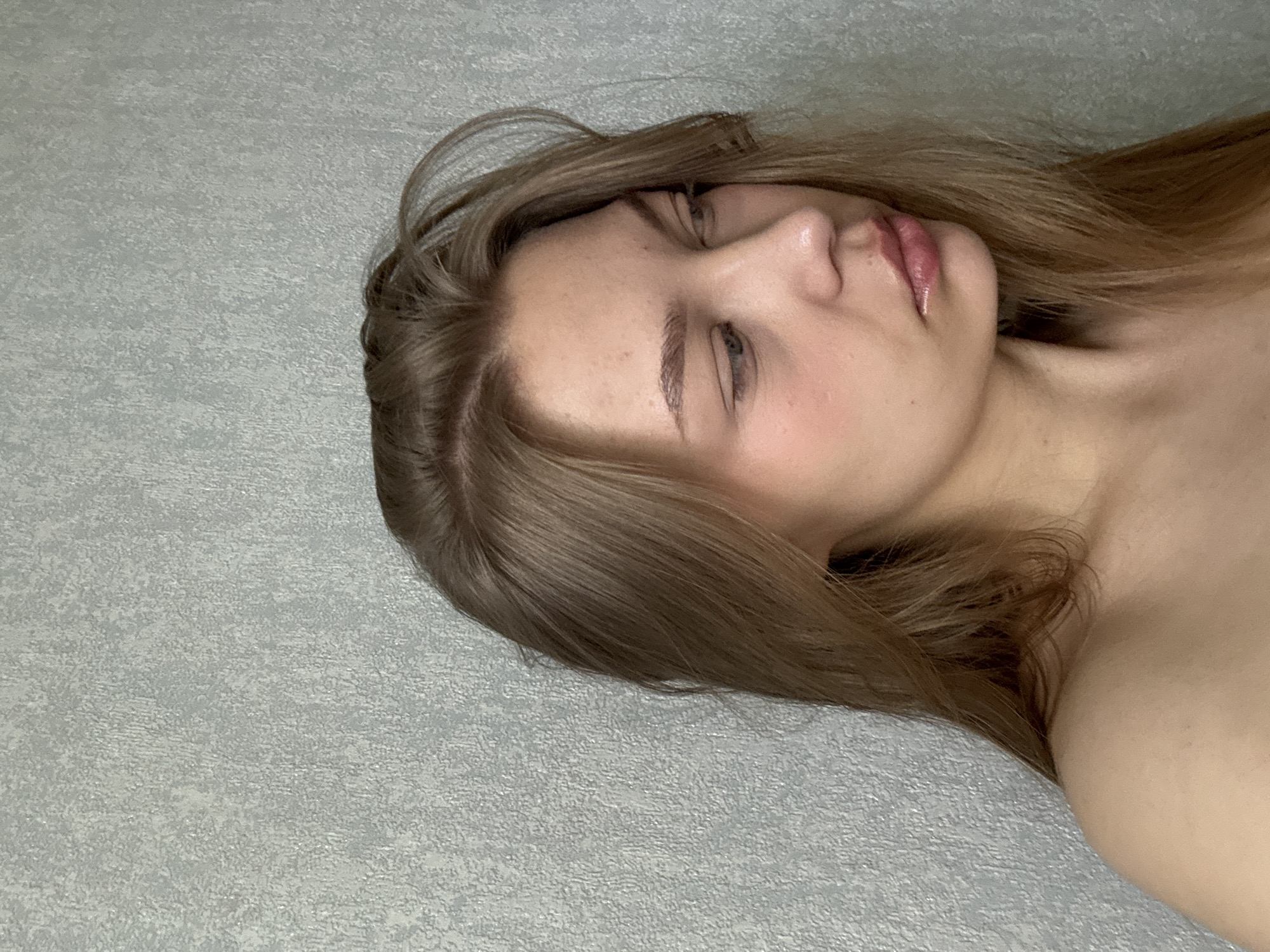 A close-up, horizontal, medium-shot image captures a young woman lying down. She has light brown hair and fair skin. Her eyes are closed, and she is slightly turned to the side. Her lips appear shiny. The background is a light gray textured surface. There is no information to confirm that the image was taken in Chaikovsky, Russia.