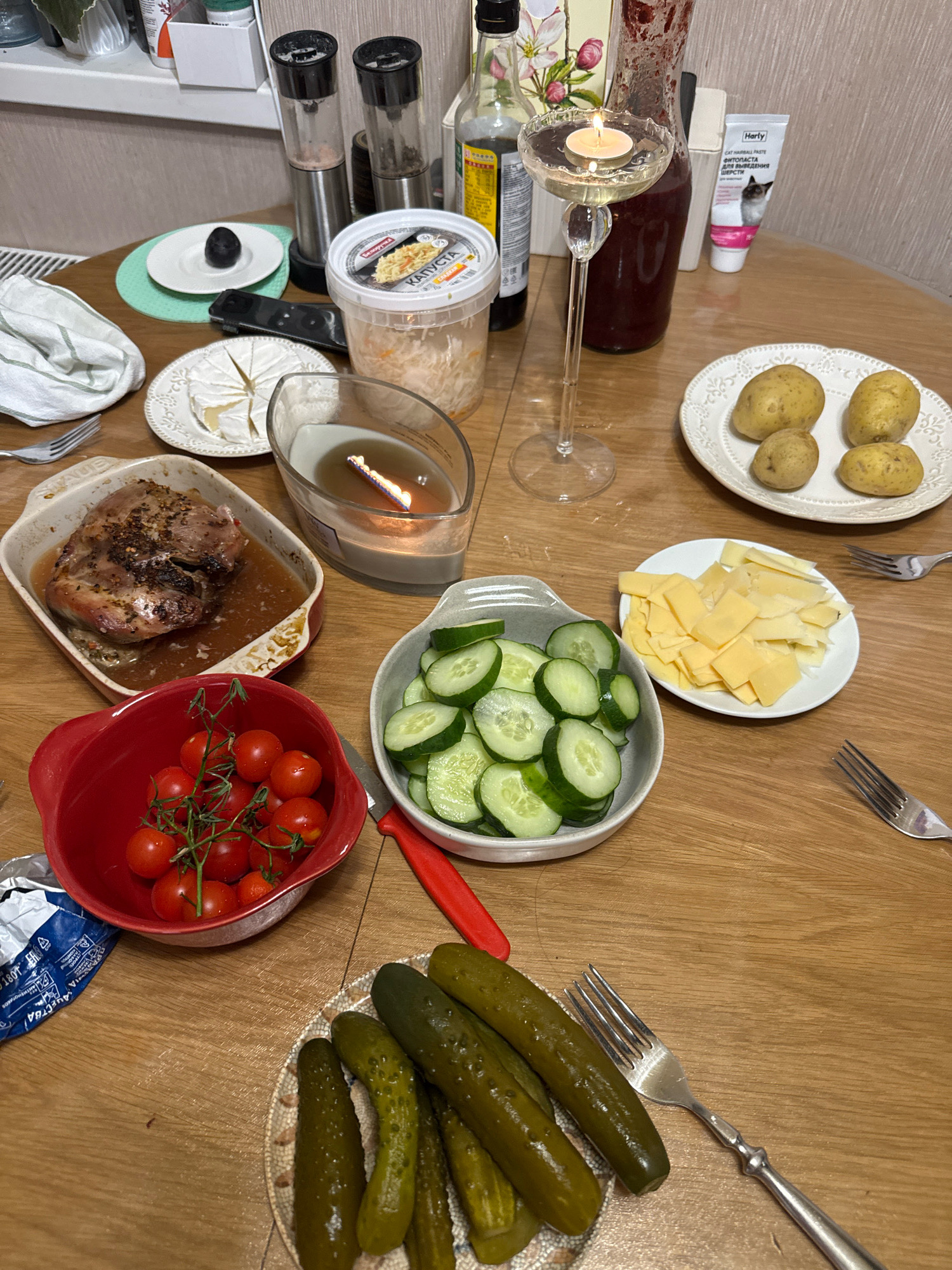 The image shows a table laden with a meal, likely in a home setting in Захарьино, Russia. A roasted piece of meat sits in a white ceramic dish, surrounded by a savory sauce. Nearby, a bowl of sliced cucumbers and a bowl of cherry tomatoes are arranged. Sliced cheese is presented on a small plate, accompanied by a portion of potatoes. Pickled cucumbers are piled on a decorative plate, with a fork resting beside them. A lit candle in a glass holder adds a warm ambiance to the scene. On a shelf in the background, various condiments and bottles are visible, including salt and pepper grinders, a bottle of soy sauce, and a container of sauerkraut. A tube of what appears to be pet care product is also present. The overall impression is of a home-cooked meal prepared for enjoyment.