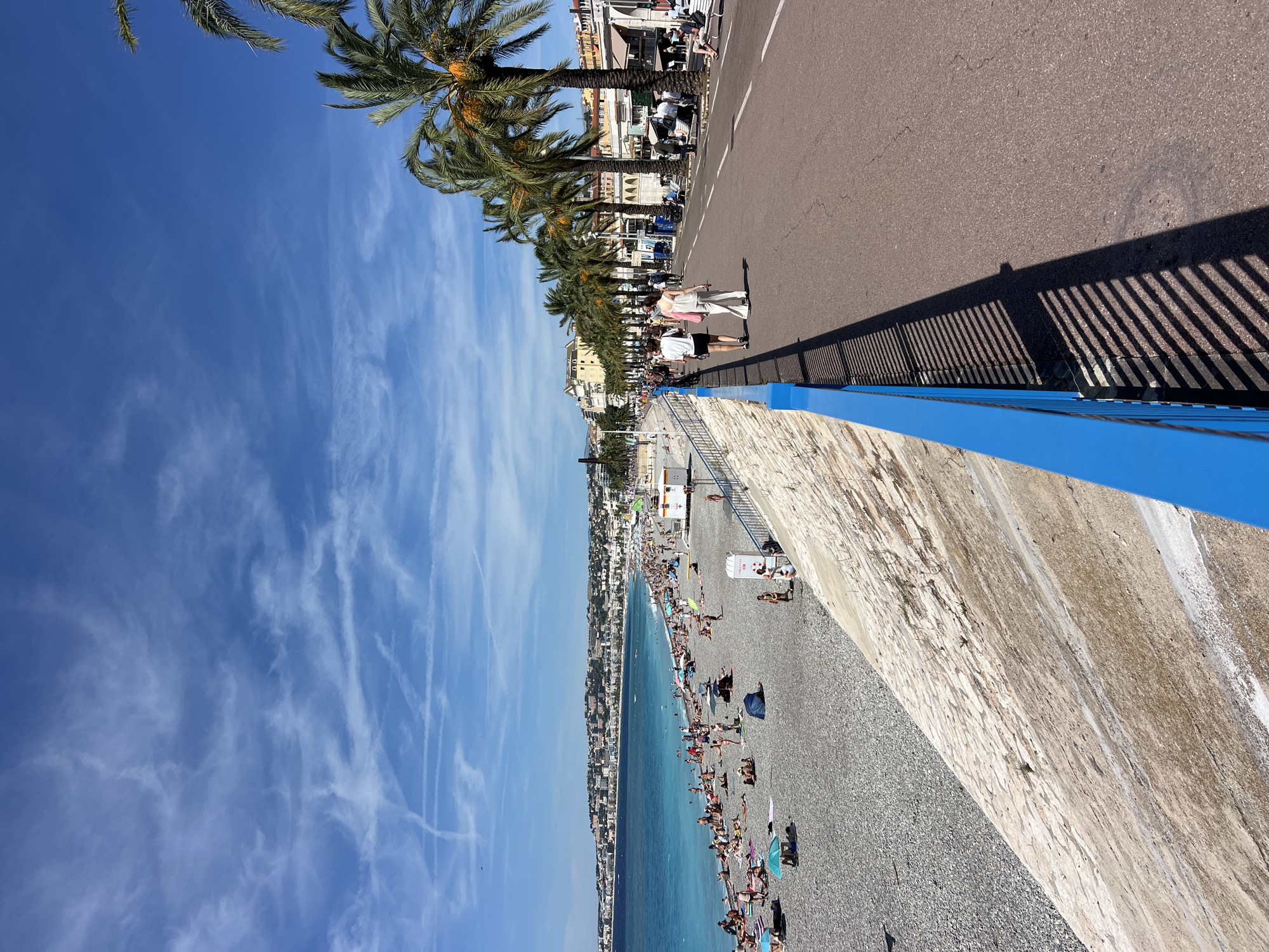 The image shows a busy pebble beach and a promenade in Nice, France, with the Mediterranean Sea in the background. On the beach, numerous people are sunbathing, some under colorful umbrellas. The water is a clear turquoise, and the coastline stretches into the distance, lined with buildings.
Adjacent to the beach is a paved promenade, bordered by a bright blue railing. Several palm trees line the promenade, casting shadows on the asphalt. People are walking and strolling along the promenade. In the background, beyond the palm trees, are buildings with light-colored facades, typical of Mediterranean architecture. The sky is blue with scattered white clouds. The scene depicts a sunny day in a popular tourist destination.