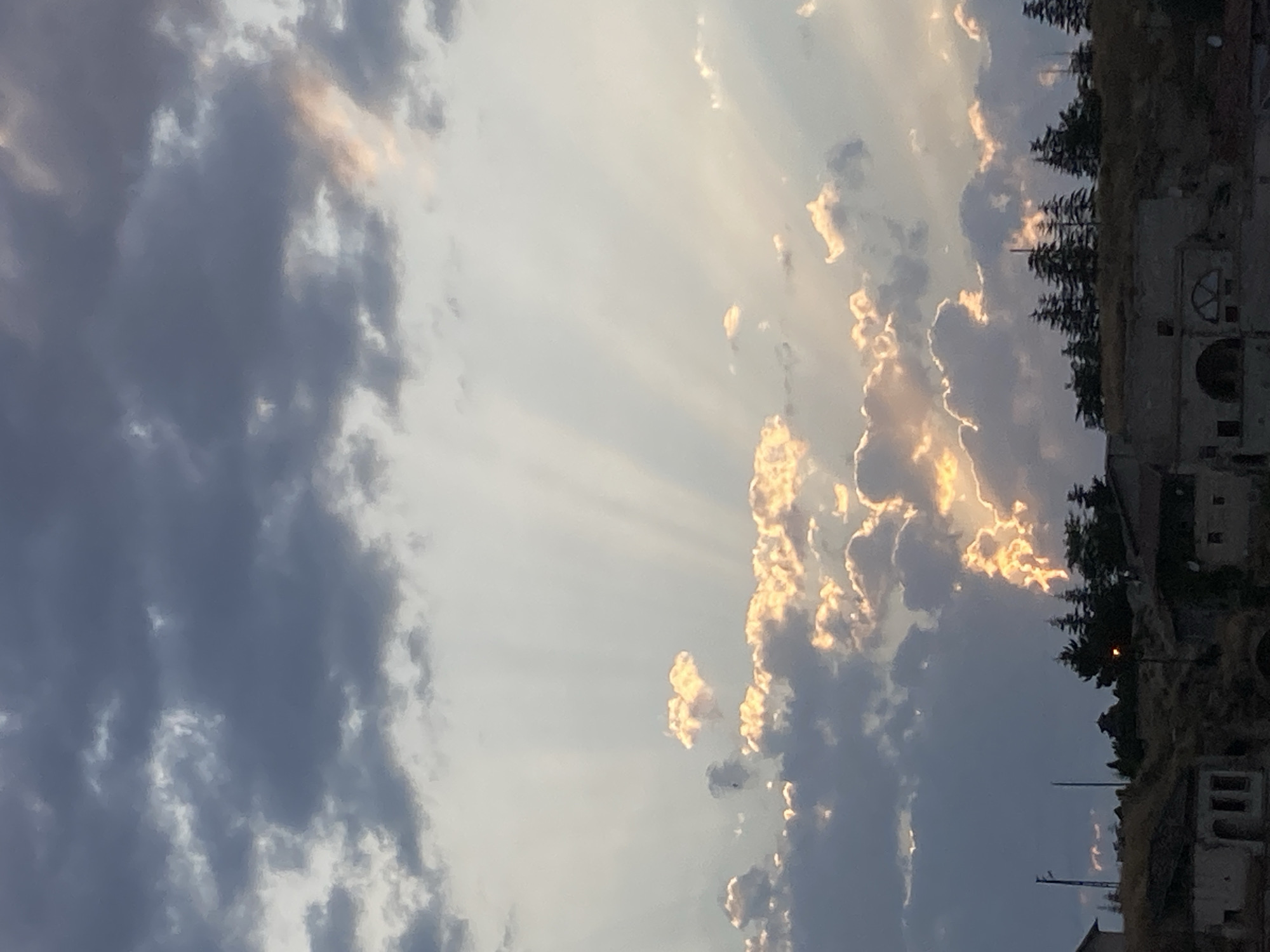 The image depicts a sky filled with dramatic clouds and sun rays over the city of Avanos İlçe Merkezi, Turkey. Dark grey clouds dominate the upper portion of the frame, contrasting with lighter, illuminated clouds that catch the golden hues of the setting or rising sun. Shafts of light emanate from behind the clouds, creating a crepuscular ray effect. In the foreground, silhouetted against the bright sky, is the landscape of Avanos, characterized by rolling hills topped with evergreen trees. At the base of the hills, traditional Turkish architecture is visible, with stone buildings featuring arched windows and rustic facades. The scene is devoid of people and activity, presenting a serene, natural landscape at dusk or dawn.