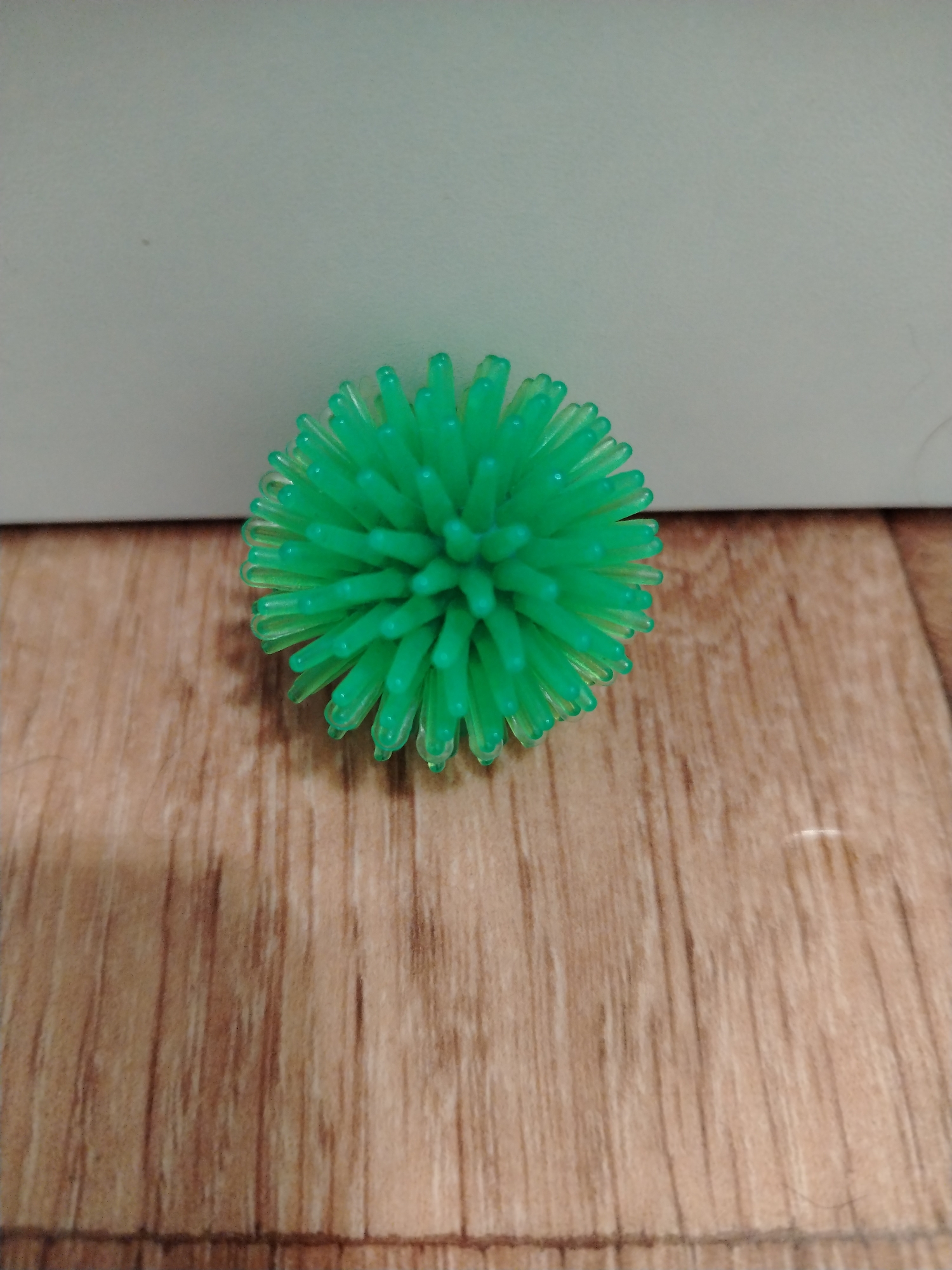 A bright green, spiky rubber ball is centered on a wooden surface. The background is a pale, muted color, possibly a wall. The ball is roughly spherical with numerous flexible, protruding spikes that give it a starburst or sea urchin-like appearance. The wooden surface has a visible grain pattern. The lighting appears to be from above and slightly to the right. The image is taken from a slightly elevated, straight-on perspective. There are no people or any other identifiable objects in the scene.