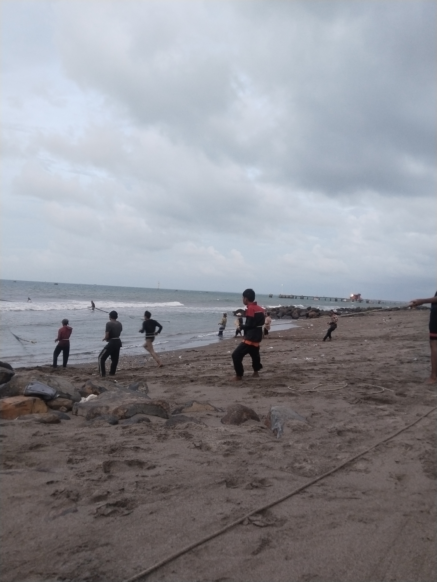 The photograph depicts a group of people working together to pull a fishing net onto the beach at Lhokseumawe, Indonesia. The scene is set on a grey sandy beach under a cloudy sky. The ocean waves are visible in the background, along with a pier-like structure extending into the water. There are about eight individuals visible, some standing on the beach holding onto a long rope connected to the net, while others are in the water near the net itself. The people appear to be engaged in a collaborative effort related to fishing. Rocks are scattered on the beach in the foreground.