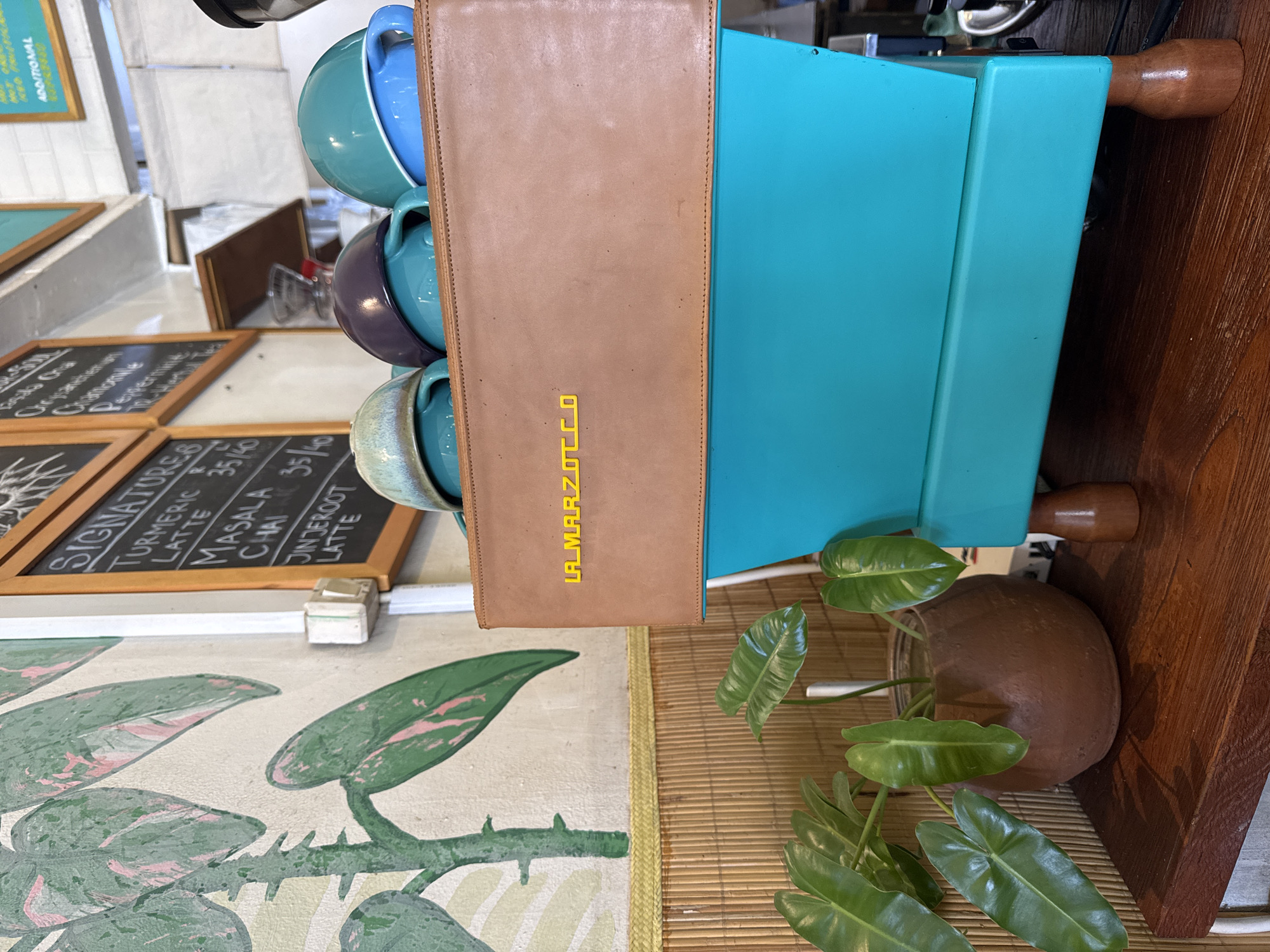 This media file captures a close-up, vertically oriented view of a vibrant cafe setting, likely located in Kuta Selatan, Indonesia. No people are visible in the frame.
The central subject is a turquoise or light blue espresso machine, featuring a tan leather-like panel on its side with the brand "LA MARZOCCO" distinctly embossed in bright yellow, block letters. A stack of ceramic mugs or bowls in varying shades of blue, teal, and purple rests on top of the machine. The espresso machine stands on dark wooden feet, suggesting it's placed on a wooden counter or shelf that extends to the right.
To the left, several black chalkboards with white handwritten text are visible. One prominent chalkboard displays "SIGNATURE" items, including "TURMERIC LATTE 35/40", "MASALA CHAI 35/40", and a partially visible "JINJEROOT LATTE". Other chalkboards show additional menu items like "CRANBERRY" and "HERBAL TEA" with some prices.
Below the counter, a patterned wall mural features large green leaves with soft pinkish accents. A light brown woven bamboo mat is laid out, upon which a small green plant in a brown pot is placed, adding a touch of nature to the scene. The overall ambiance suggests a welcoming and relaxed environment typical of a modern cafe or coffee shop, likely in operation during the daytime given the bright lighting. The prices listed on the chalkboards (e.g., 35/40) likely refer to local currency.