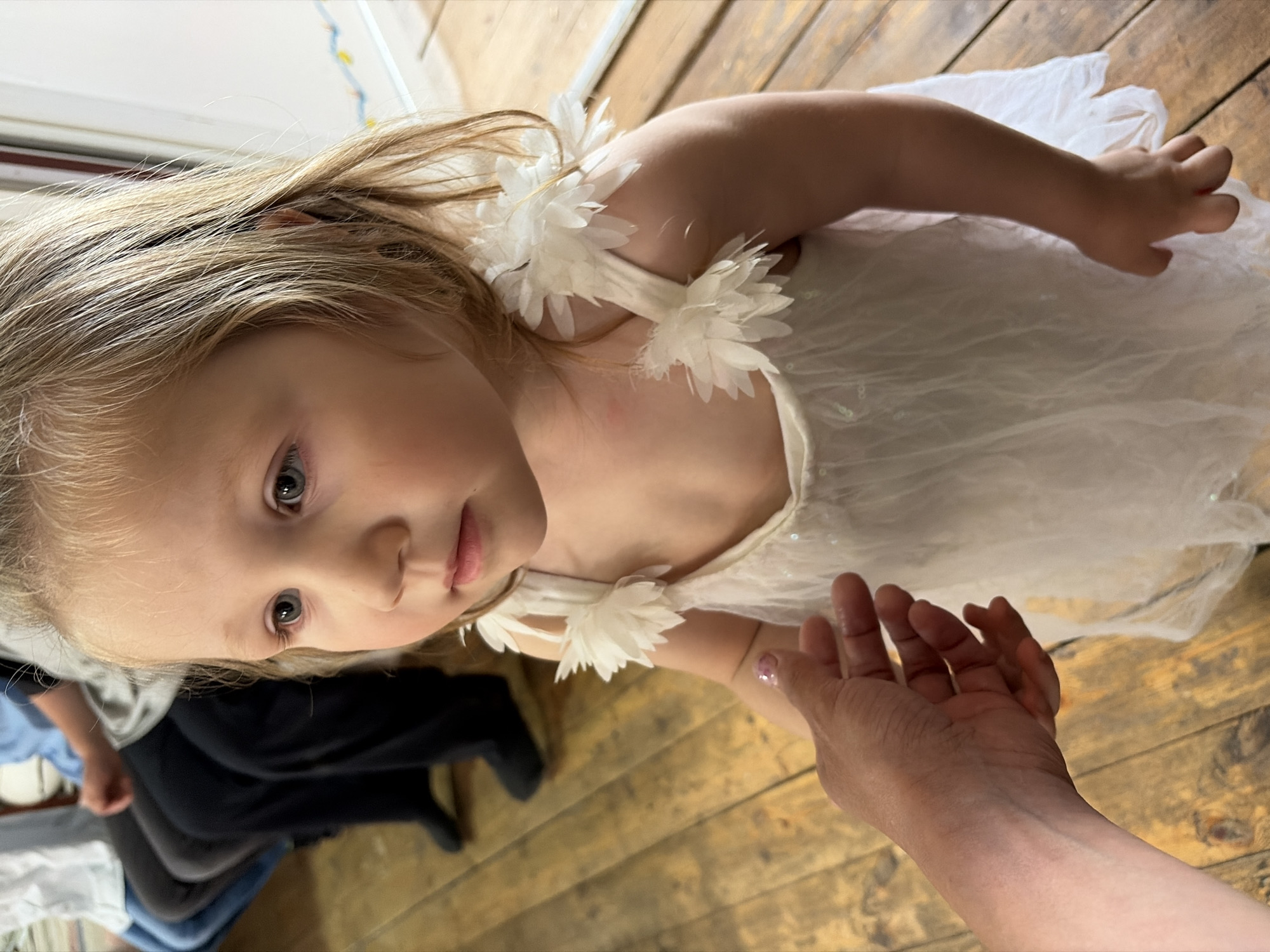 A young child with blonde hair and striking blue eyes is the central focus of this indoor portrait, captured from a slightly elevated, downward-looking perspective. The child is looking upwards towards the camera with a calm, inquisitive expression. She is wearing a delicate white dress featuring decorative, layered floral or feather-like elements on the shoulder straps, suggesting a special occasion or playful attire.
An adult's hand and forearm are prominently featured in the lower right foreground, reaching towards the child, implying an interaction, perhaps an offer of support or engagement. The hand has a subtle sheen, possibly from lotion or a very light, clear nail polish.
The background primarily consists of a warm-toned wooden floor with visible grain, suggesting a rustic or home-like setting. Blurred forms of another person's clothing, including dark trousers and a lighter garment, are partially visible in the left background. The lighting is soft and even, consistent with an indoor environment. No specific time of day is discernible, but the scene is well-lit. There is no visible text. The image conveys a tender, candid moment of interaction within a cozy indoor space in Прикамский, Russia.