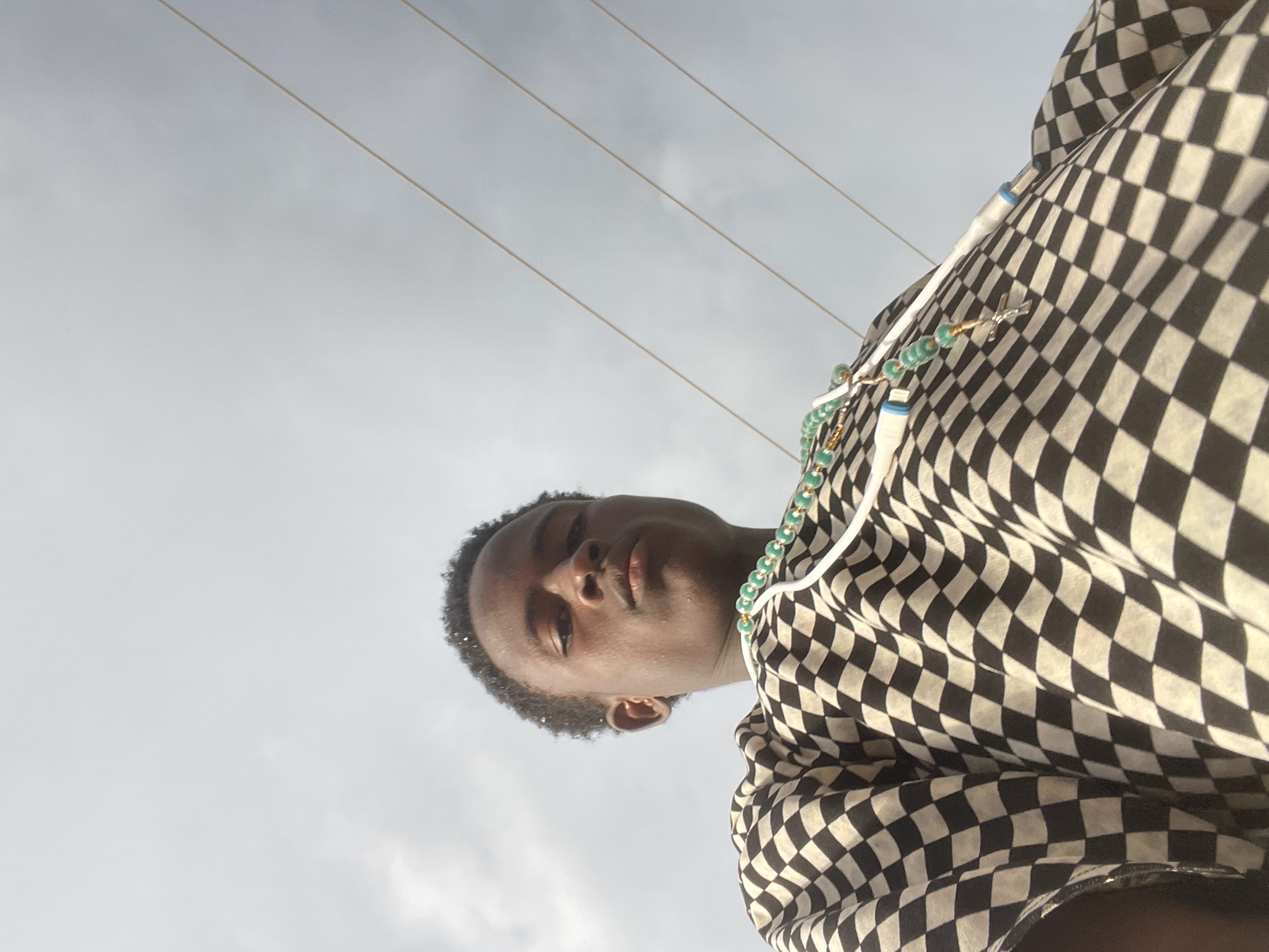 The image shows a low-angle, waist-up shot of a young Black male facing the camera. He has dark skin and short, dark curly hair. He is wearing a black and white checkered shirt and a beaded necklace with green beads and a silver cross pendant. The background is a pale, cloudy sky with three thin, light-colored power lines stretching diagonally across the frame. The image appears to be taken outdoors during the day in a location that could be the city of Jos, Nigeria, given the context. There is no clear indication of specific urban landmarks to pinpoint the exact location within Jos. The overall lighting suggests an overcast day.