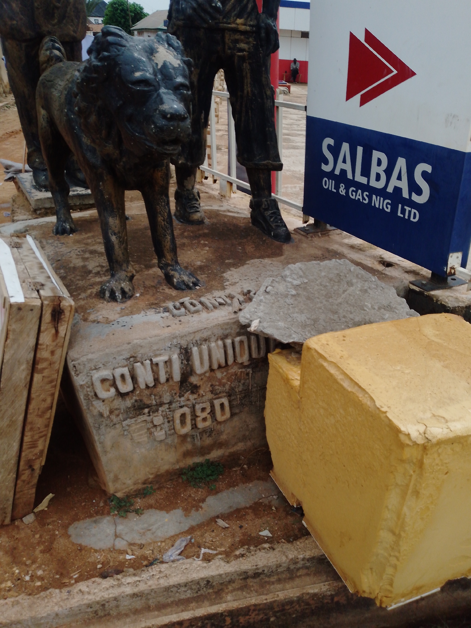 The scene captures a portion of a statue in Sabon Tasha, Nigeria. A bronze sculpture of a dog stands next to human figures. A sign for SALBAS Oil & Gas Nig LTD is visible in the background. Concrete blocks and wooden planks surround the statue's base, which has the inscription "CONTI UNIOU 080". The ground around the statue is dusty and dirty. A lone individual is faintly visible in the background near the SALBAS sign.