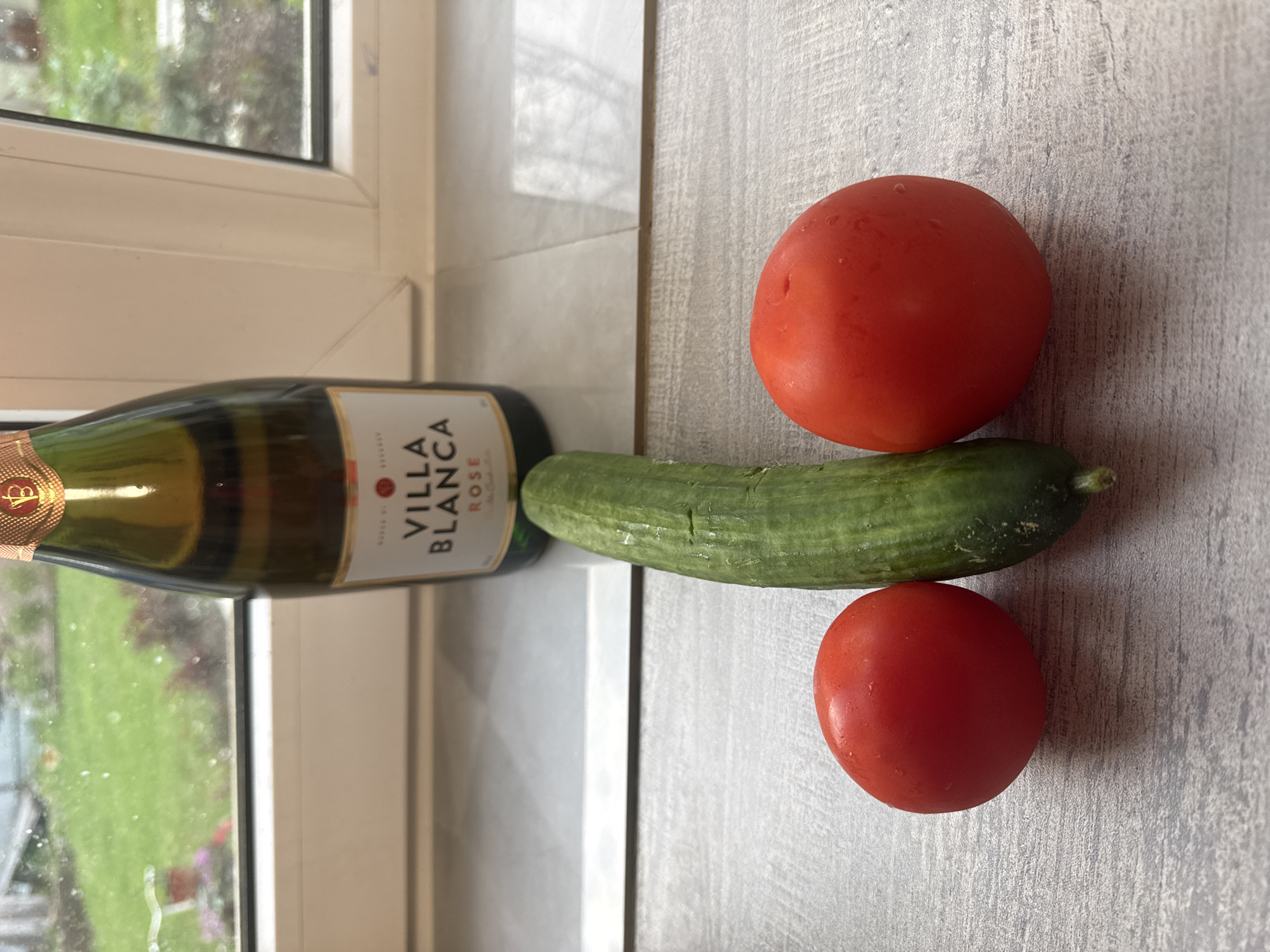 A horizontal, medium-shot photograph depicts a bottle of Villa Blanca Rose, a cucumber, and two tomatoes lying on a surface. The bottle is positioned on its side near a window, with its label reading "Villa Blanca Rose." A green cucumber runs horizontally across the center of the frame. A tomato is located on each side of the cucumber. The surface underneath the objects appears to be a light gray countertop. The window shows a view of foliage outside. There is no indication of the city of Chaikovsky, Russia. There are no people in the photograph.