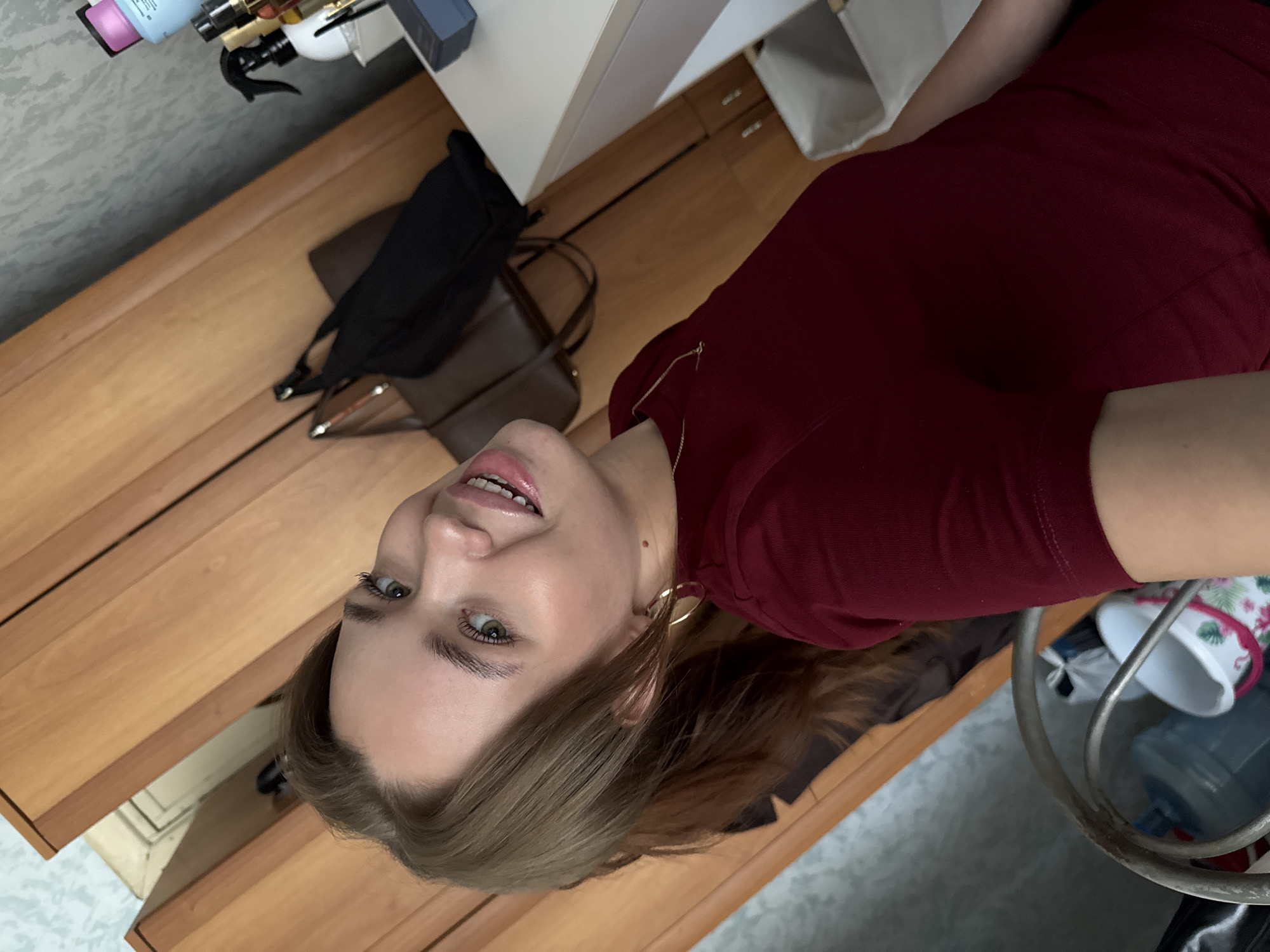 The image shows a young woman with light brown hair and green eyes, wearing a maroon t-shirt and a gold necklace. She is indoors, in what appears to be a bedroom or dressing area. The background includes a wooden wardrobe, a white dresser with various cosmetic products, and a basket containing what looks like plastic bags and a floral-patterned container. A large water dispenser can be seen in the lower left corner. The lighting is natural, coming from a window or overhead source. The woman is looking towards the camera with a slight smile.