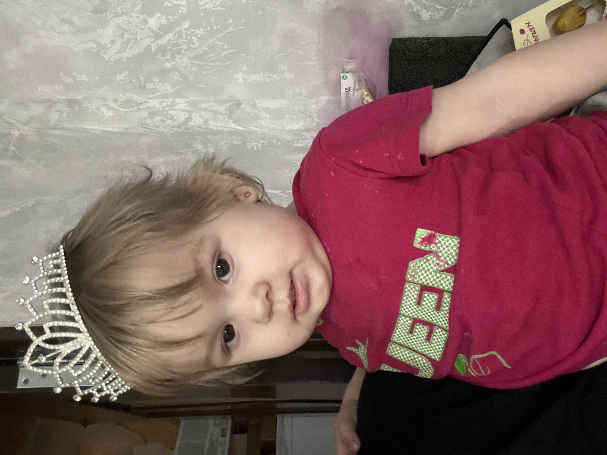 A young child with light brown hair is wearing a silver tiara. They have brown eyes and are wearing a pink t-shirt with green lettering. The child is lying down. The background includes a textured white curtain, a package of what appear to be medical supplies, and a black textured object. It is not possible to determine if the image was taken in Ольховка, Russia, from the given information.