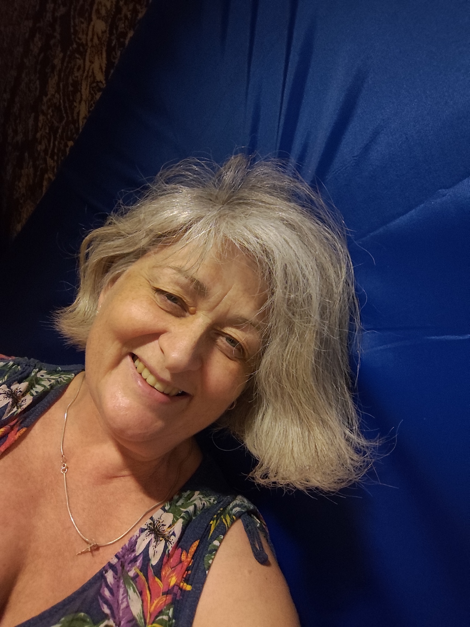 The image is a selfie of a middle-aged woman with graying blonde hair and light skin. She is smiling and looking at the camera. She is wearing a floral print top and a silver necklace. The background is a blue fabric that appears to be a curtain or a bedspread, with a patterned fabric visible in the upper left corner. The woman's head and upper body are visible in the frame. The lighting suggests an indoor setting. The image does not contain any information that would indicate its location in Krasnodar, Russia.