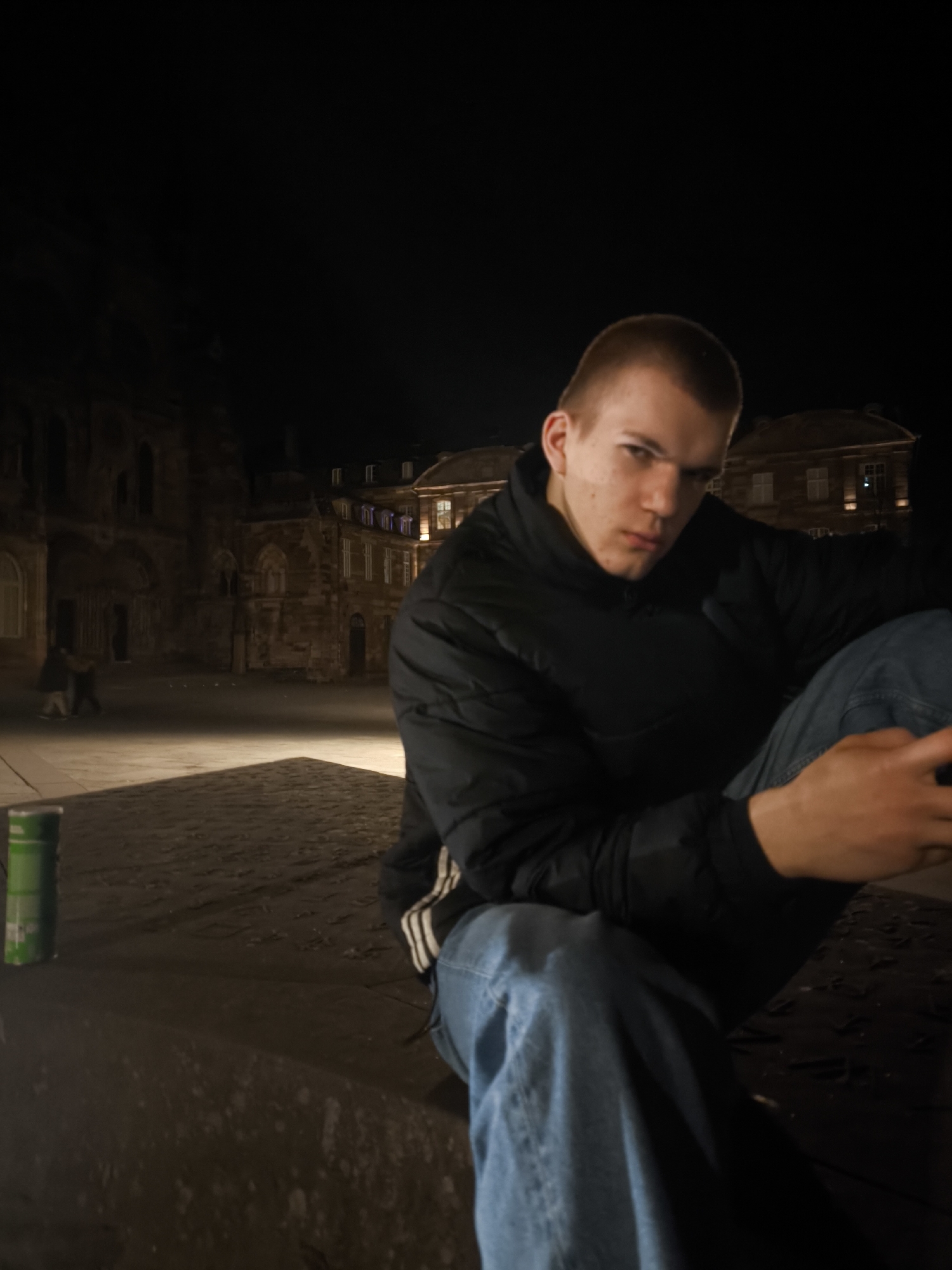 The image depicts a young man sitting in an outdoor urban setting at night. He is wearing blue jeans and a black jacket with three white stripes on the sleeve. A green can is visible to the left of the frame. In the background, there are buildings that appear to be located in Strasbourg, France, with architectural details consistent with buildings in the city. Two individuals can be seen walking together in the background. The area is lit by artificial light, creating shadows and highlighting the cobblestone ground.