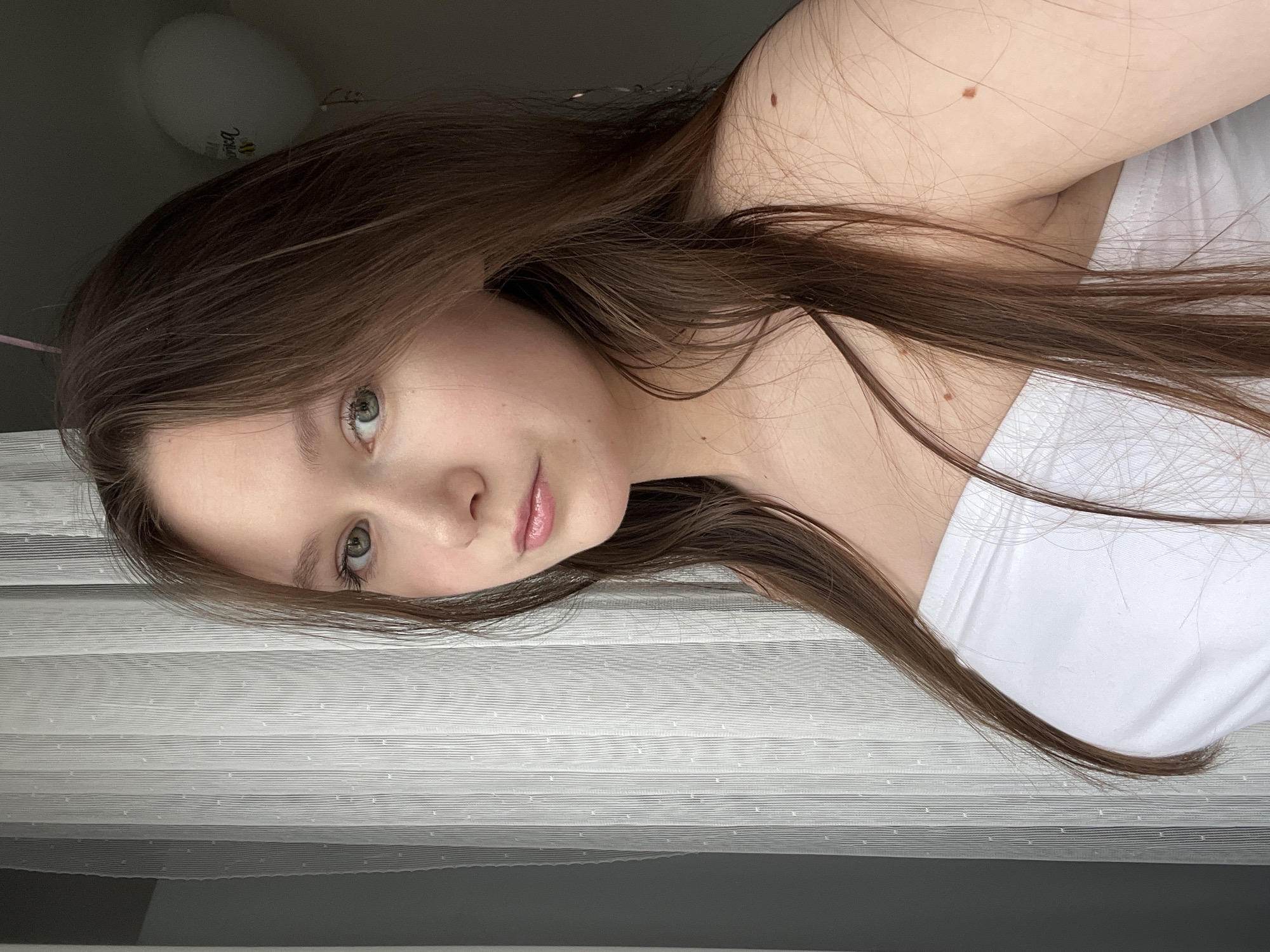A close-up, rotated portrait-style image features a young individual, likely female, with long, straight brown hair, green eyes, and fair skin. The person is looking directly at the camera with a neutral to slightly smiling expression. They are wearing a white strapless top or undershirt.
The image appears to be a selfie taken indoors during the daytime, evidenced by the soft, natural light. In the background, to the left (when oriented correctly), a white balloon is visible. The balloon displays a crown logo and the word "Corona," with additional fainter text that appears to say "Happy Birthday" and "Extra," suggesting a branded celebratory balloon.
The lower half of the image (when oriented correctly) reveals textured white window blinds or curtains, indicating a room with a window. Several small, brown moles or freckles are visible on the person's shoulder and upper arm. The overall mood is calm and natural. The location context for this individual is Chaikovsky, Russia.