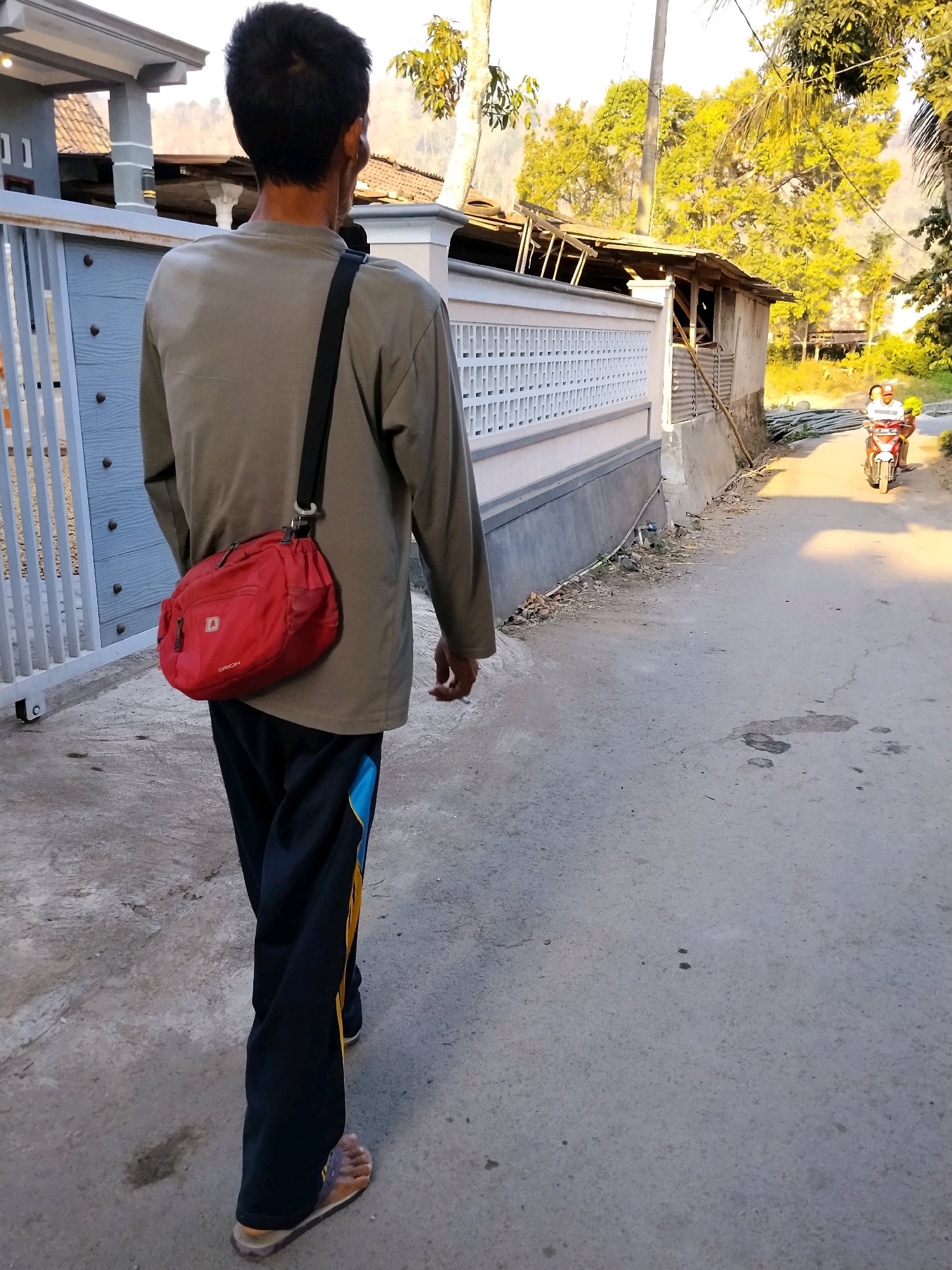 A person is walking away from the camera on a paved road in Tlogosari, Indonesia. The person is wearing a long-sleeved, olive-green shirt, black pants with yellow and blue stripes, and sandals. A red messenger bag is slung over their right shoulder. In the background, a white fence borders a house, and further down the road, a person on a red motorcycle is visible. Trees and foliage are present on the right side of the image. The scene appears to be a residential street with some construction or dilapidated structures visible.