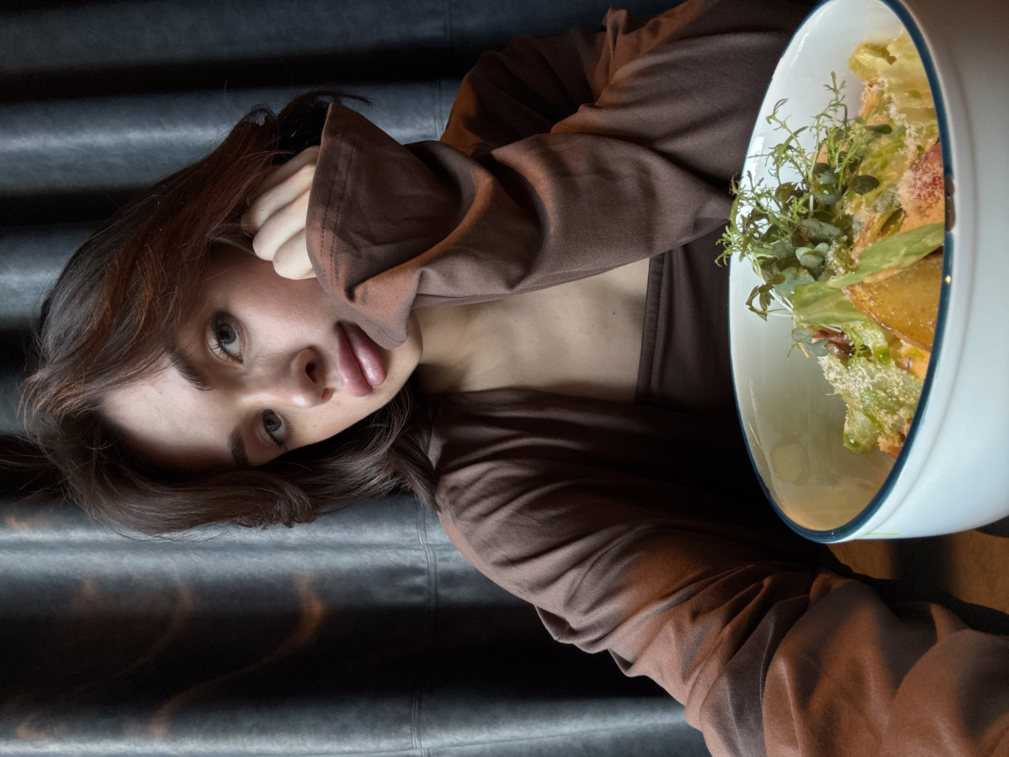 A young woman with dark hair and fair skin lies on a gray, textured surface, possibly a couch or bed, next to a large white bowl filled with a salad. She wears a brown outfit. She's holding her hand up to her face and slightly sticking out her tongue. The salad bowl is primarily filled with green lettuce and herbs, and also contains fried bread and shredded cheese. The location is unknown.