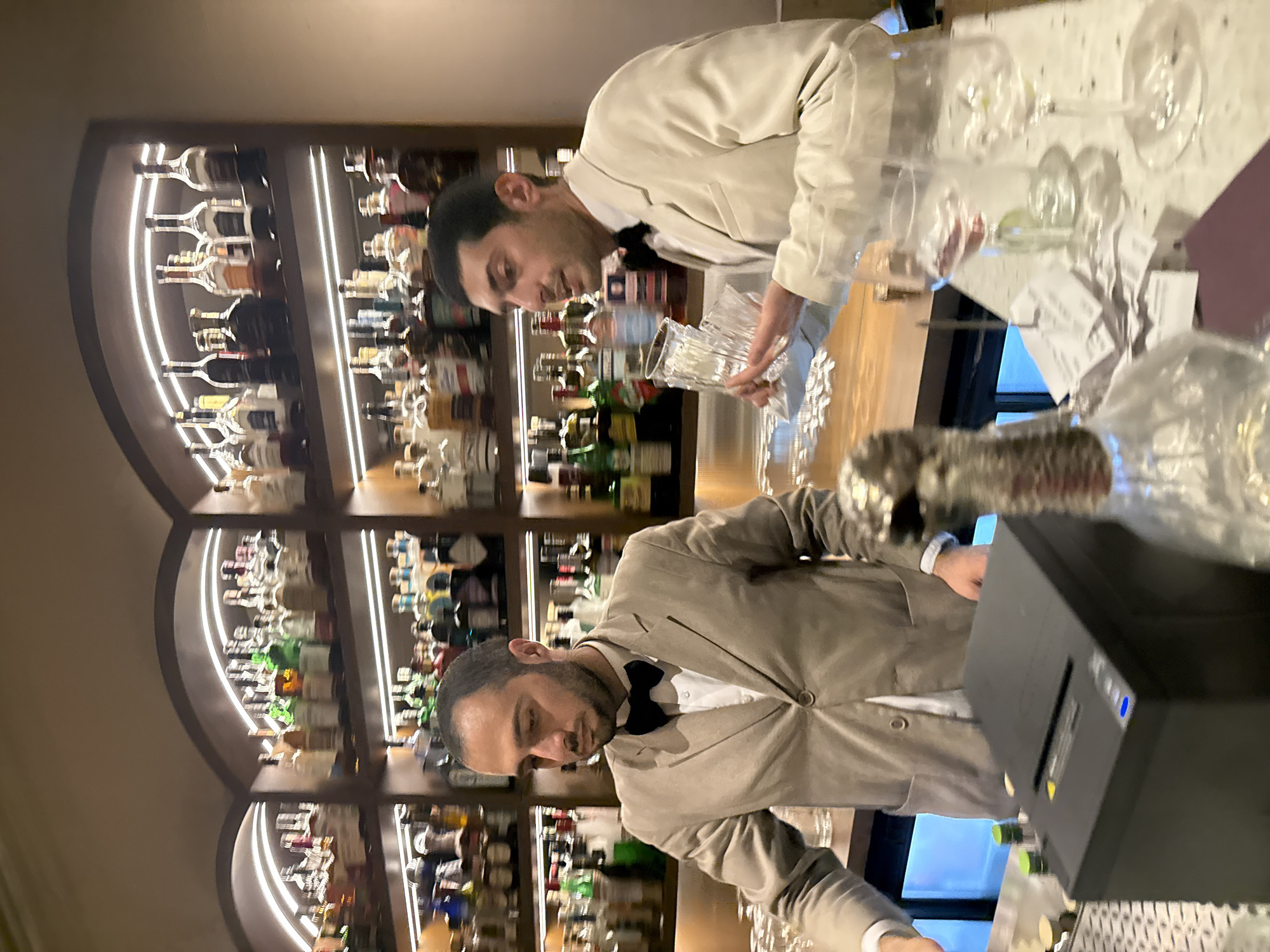 A photograph shows two men behind a bar. The bar has a large shelf lined with bottles of alcohol. The men both wear light-colored jackets with bowties and white shirts. One man holds a glass decanter. The other man stands next to a black rectangular object, possibly a cash register or printer. The setting appears to be a bar. The location is Yerevan, Armenia.