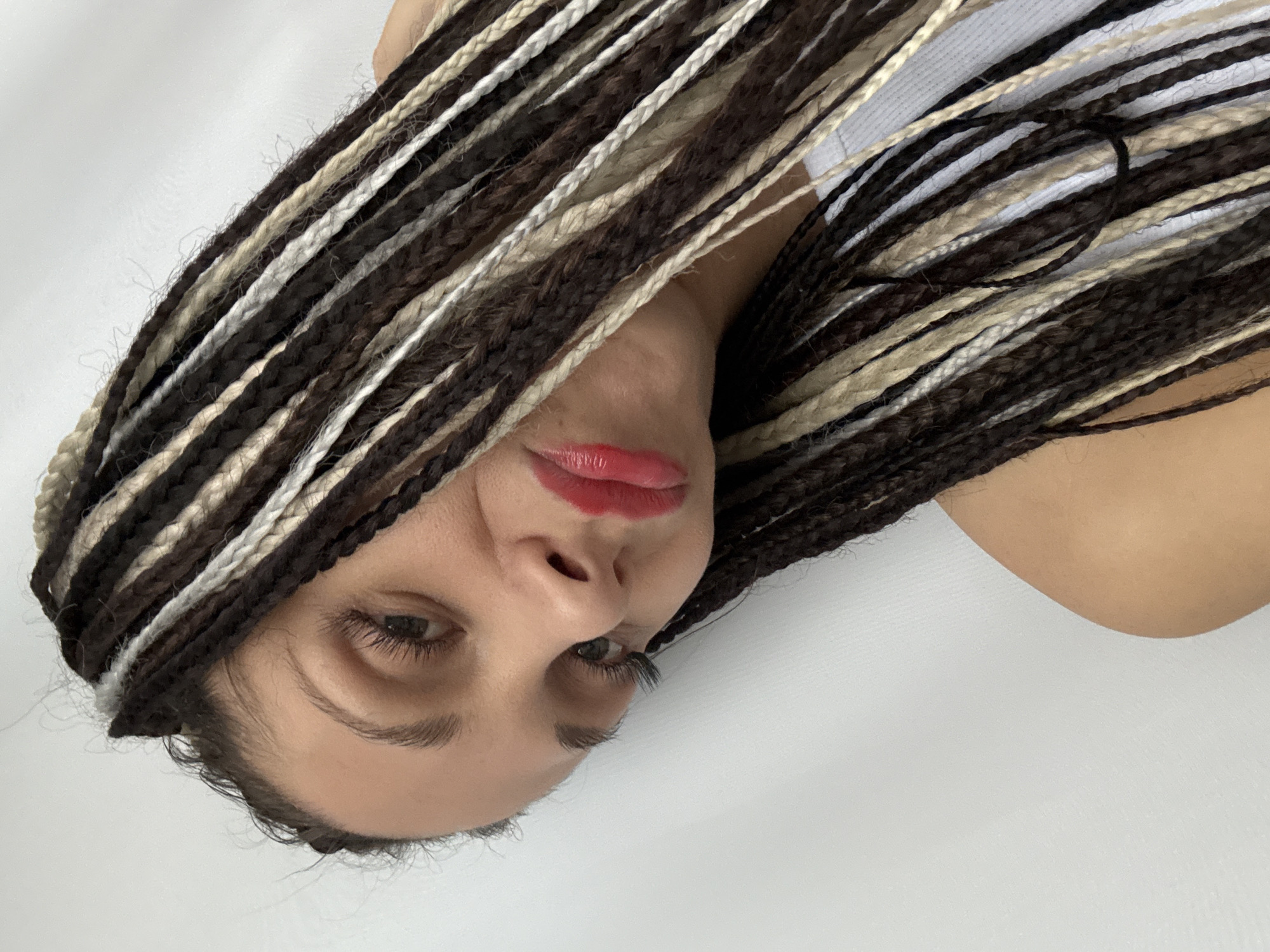 The image is a close-up, tilted portrait of a person with dark brown hair braided with beige and white strands. The person has fair skin, brown eyes, and is wearing red lipstick. A portion of a white top is visible in the background. The background is a plain white surface. The location is not determinable from the image content. The specific city is unverifiable from the visible elements.