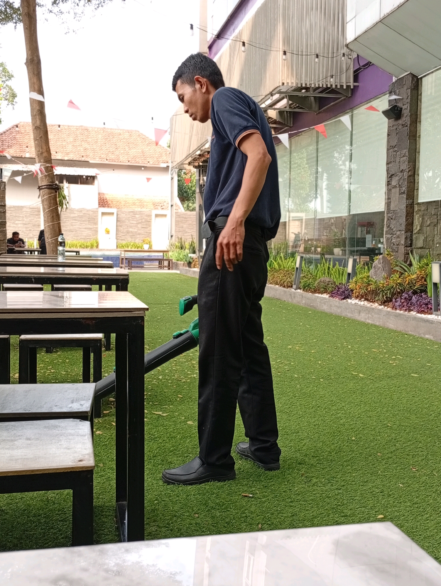 A man in a dark blue short-sleeved polo shirt and black trousers is standing on a green artificial turf surface in front of a row of outdoor tables and chairs. He is holding a green and black leaf blower, which is angled downwards towards the turf. The man is looking down and to his left. Behind him, there is a building with large glass windows and a textured stone facade. Red and white flags are visible strung across the facade. In the background, there are more trees and buildings, with a person seated at one of the tables. The scene appears to be an outdoor dining or seating area.