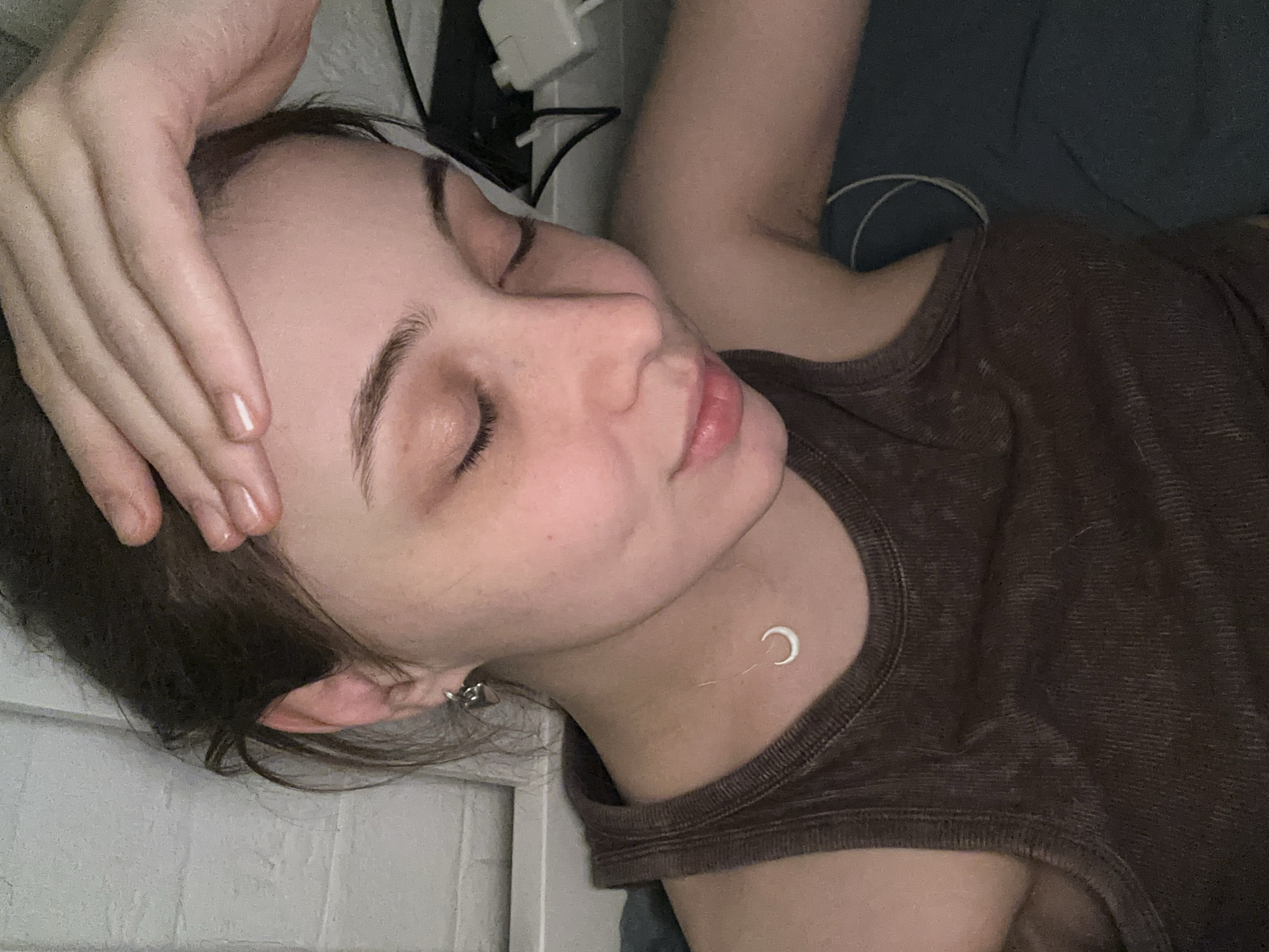 A young woman is lying down, with her hand resting on her forehead. She has fair skin, dark hair, and is wearing a brown tank top and a silver necklace with a crescent moon pendant. She is also wearing earrings. The background shows what appears to be a wall and bedding. The location is not discernible from the image.