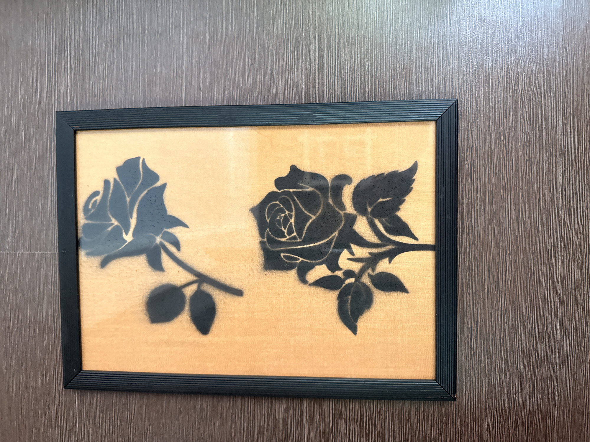 A framed artwork featuring two black rose silhouettes on an orange background is displayed on a wooden surface. The frame is black and has a ribbed texture. The background appears to be a woven fabric. The artwork is positioned horizontally within the frame. The wooden surface has a grain pattern and is dark brown in color. The scene is indoors.