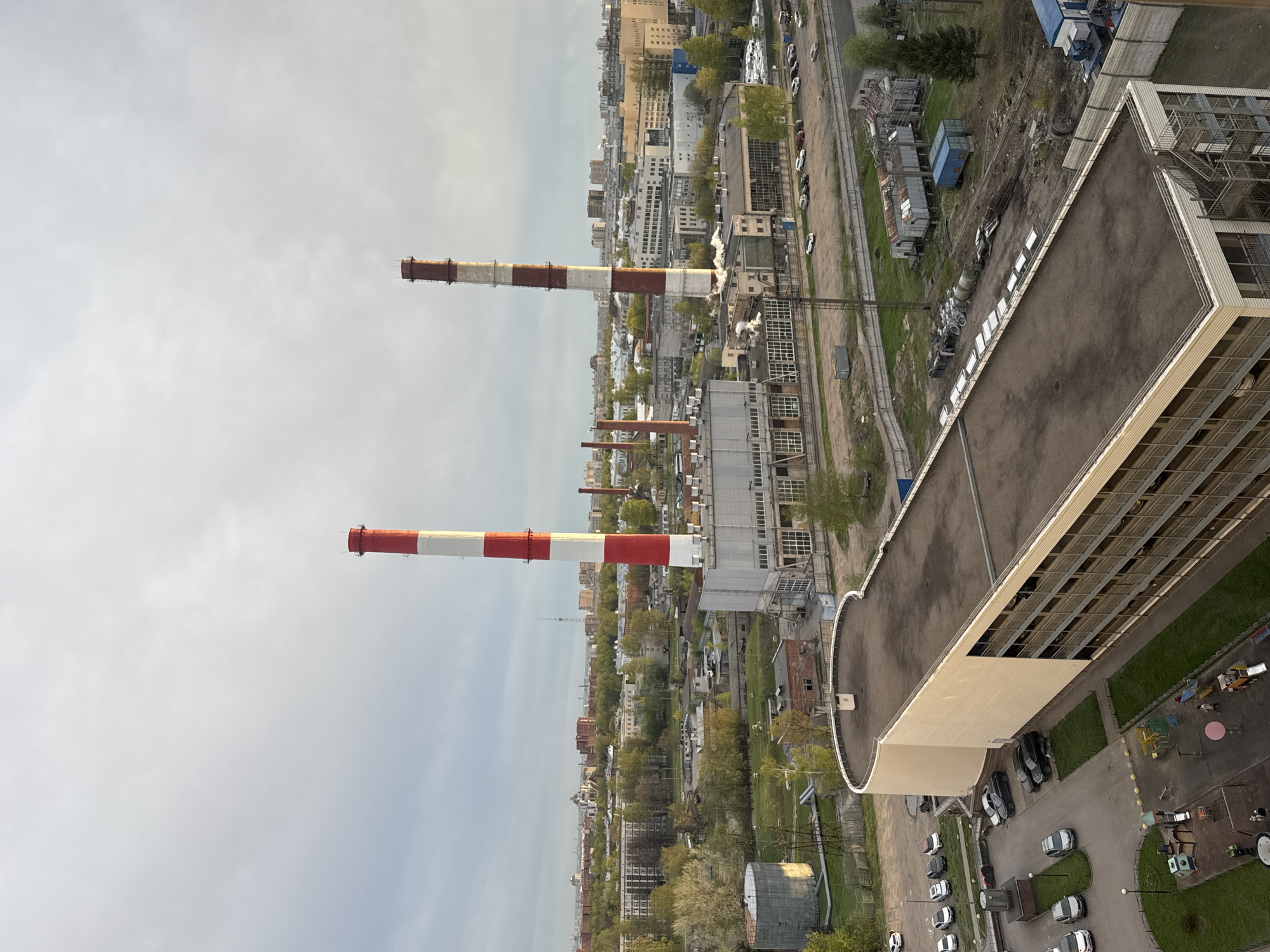 The image shows a high-angle view over a cityscape, likely in an industrial area of Unknown, Russia. The sky is overcast. Several tall, red and white striped smokestacks dominate the scene, suggesting a power plant or factory complex. The surrounding environment includes various industrial buildings, warehouses, and residential buildings further in the background. Vegetation like trees are visible between the structures. In the foreground, the edge of a building with windows and a flat roof is present. A parking lot and a small playground are visible at the edge of the image. The buildings are generally grey and beige, indicating a somewhat drab urban landscape. There is no visible human activity in the immediate view.