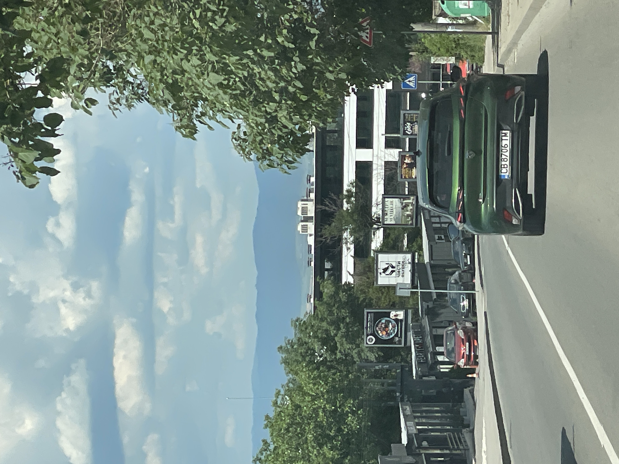 The image shows a street scene in Sofia, Bulgaria, on a sunny day with scattered clouds. In the foreground, a green Peugeot 3008 car with license plate CB 8706 TM is driving on a paved road. The car is in motion, and its taillights are illuminated. The road has lane markings and a white dashed line.
To the left of the road, there are several parked cars, including a red Renault Clio. There are also several storefronts and businesses with outdoor seating areas. Several billboards are visible, displaying advertisements. One billboard features an image of a meal, another shows a silhouette of a dog and text in Bulgarian, and a third shows a graphic of a black shoe with text in Bulgarian.
In the background, a modern, multi-story building with a white facade and dark-tinted windows is visible. Behind the building, distant blue mountains can be seen, suggesting a hilly or mountainous terrain surrounding the city. Green trees and foliage are present throughout the scene, framing the view and adding to the urban greenery. A street sign with a blue pedestrian crossing symbol and a red and white triangular warning sign are also visible on the right side of the road. A green garbage bin is also present on the right sidewalk. The overall impression is a typical urban street in a city with a mix of modern architecture, older establishments, and natural elements.