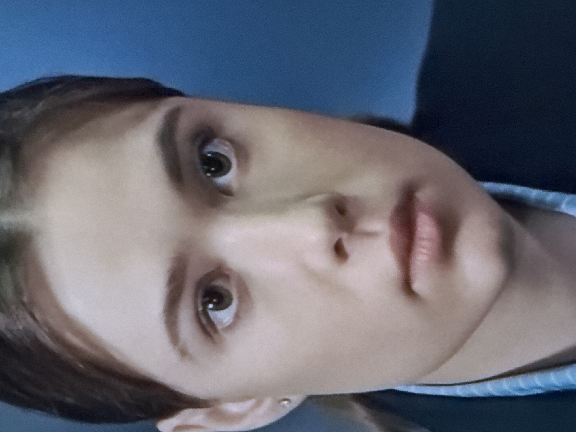 A close-up, head and shoulders shot shows the upper body of a young woman with light brown hair and fair skin, facing forward against a blurred blue background. She has dark brown eyes, defined eyebrows, and a neutral expression. She is wearing a dark blue blazer over a light blue striped shirt. A small, silver stud earring is visible in her left ear. The image is slightly grainy.