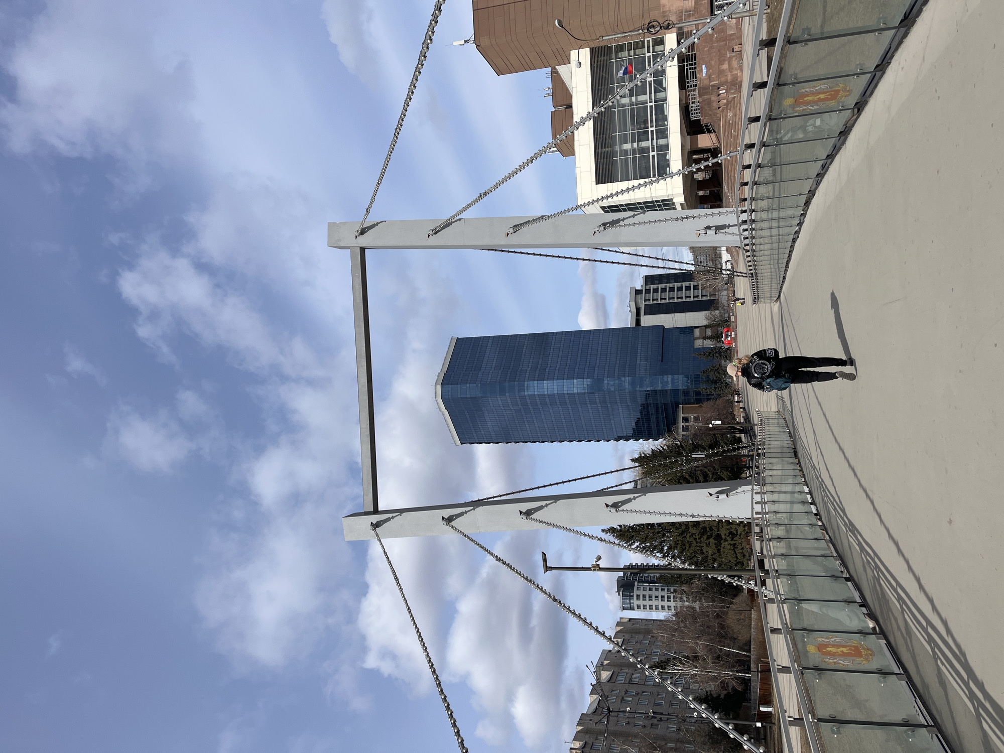 The image shows a person walking on the Vinogradovsky Bridge in Krasnoyarsk, Russia. The bridge is a pedestrian bridge with a modern design, featuring white support structures with chain supports. The walkway is concrete, bordered by glass panels with ornate emblems. In the background, a modern blue skyscraper is visible. Other buildings and trees are interspersed in the urban landscape. The sky is light blue with scattered clouds. The person is walking away from the camera, casting a shadow on the bridge surface.