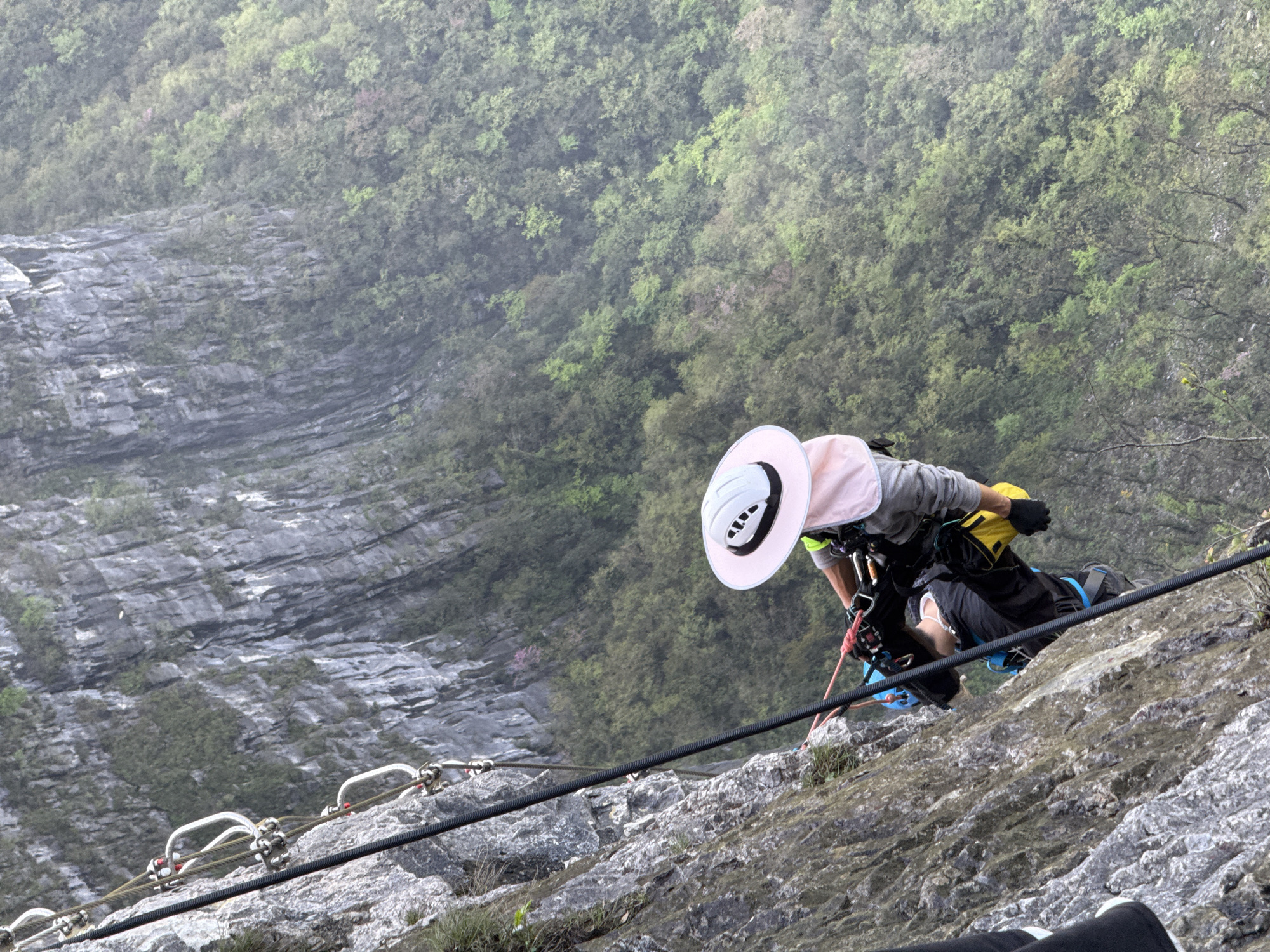 A person wearing a helmet and pink-brimmed hat is rappelling down a steep, rocky cliff face. The person is equipped with a harness and other climbing gear, and is using a rope secured to a series of metal anchors. The background shows a lush, green mountain landscape obscured by mist, and a steep drop below. This scene is likely located on a via ferrata, in the Yongding District, China.