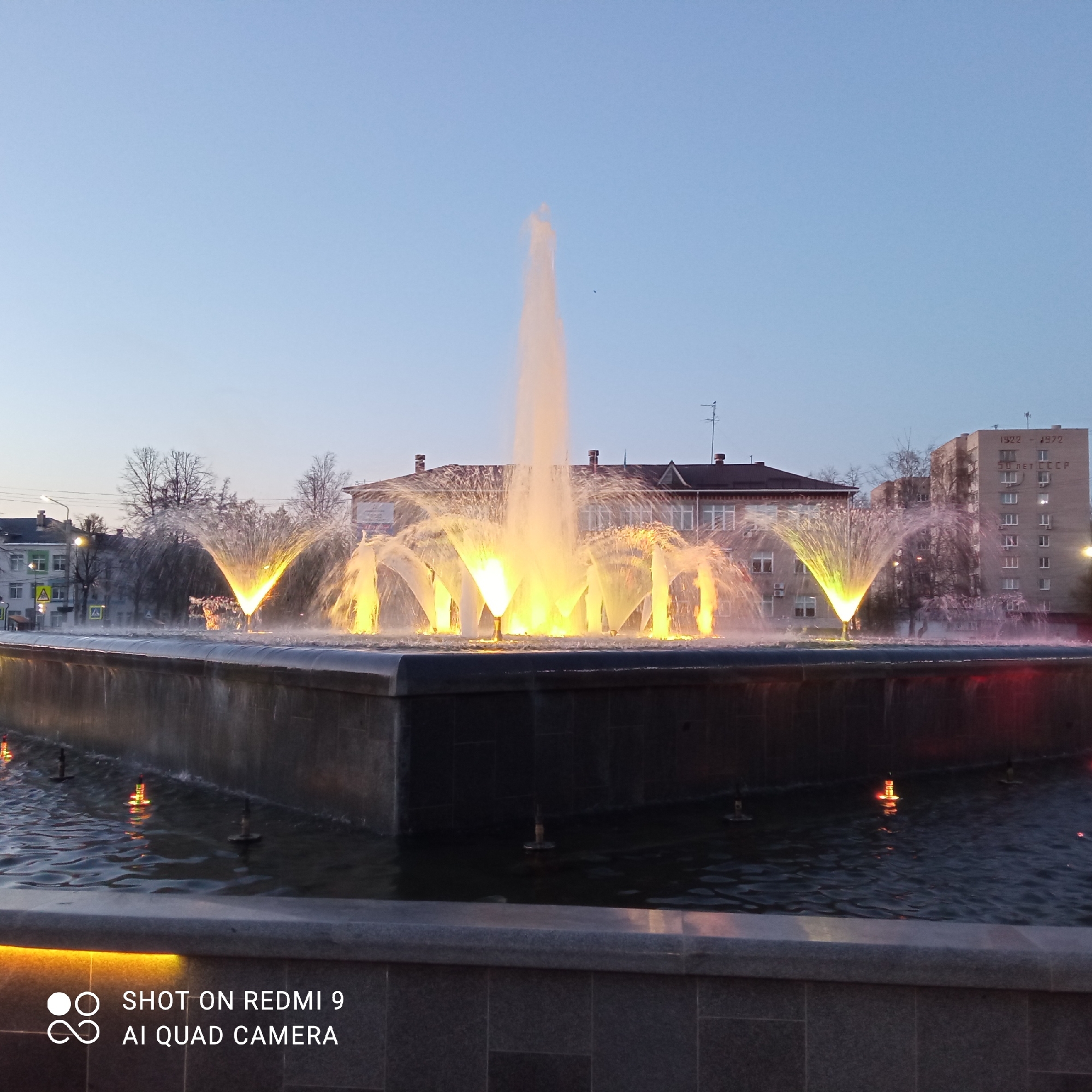 Here is a description of the image:
The image depicts a fountain in the city of Chaikovsky, Russia. The fountain features a central jet of water shooting upwards and several smaller jets arranged around it, creating an illuminated display of water formations. Buildings can be seen in the background, including one with the inscription "1922-1972 50 LET CCCP" which translates to "50 Years of the USSR." The scene is illuminated with artificial lights, suggesting it is dusk or night. The fountain's surrounding structures appear to be made of stone or concrete. There are trees without leaves in the background indicating it is either autumn or winter.