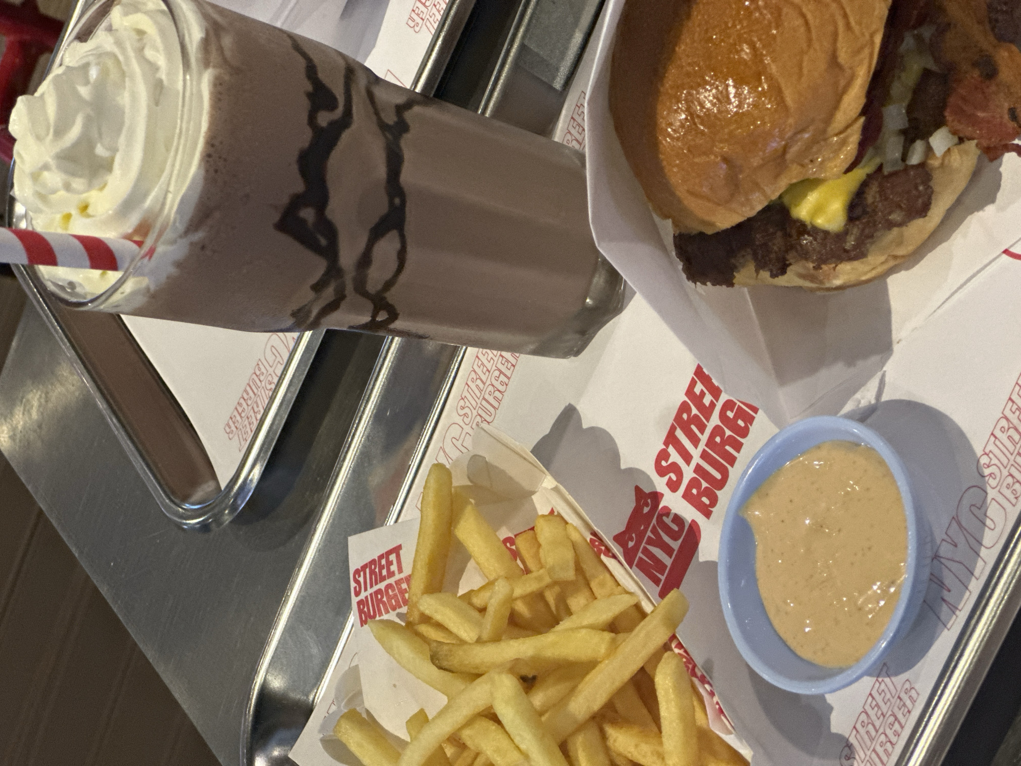 The image shows a meal from NYC Street Burger in Baan Chaweng, Thailand. The meal consists of a chocolate milkshake with whipped cream and a red and white striped straw, a burger in a white paper container, french fries in a paper container, and a small light blue bowl containing dipping sauce. The food is arranged on metal trays with the "NYC STREET BURGER" logo printed on the white paper underneath the food items.