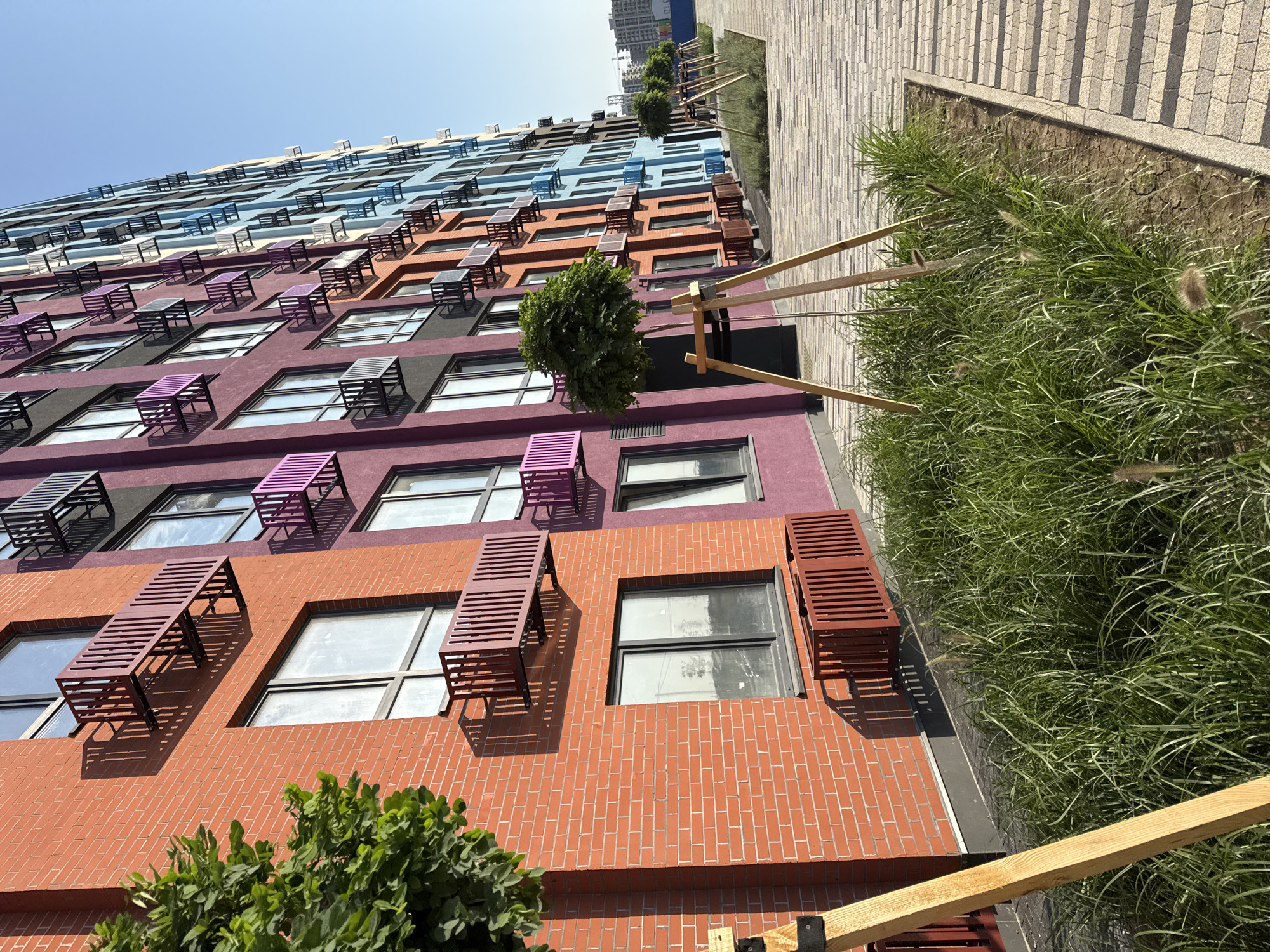 The image shows a brightly colored, multi-story residential building with a row of trees and landscaping along a paved walkway. The building features alternating sections of orange, purple, and blue facades, each with numerous windows and small metal balconies or air conditioning units. The walkway is paved with light-colored interlocking stones, bordered by a strip of green grass and small, decorative trees supported by wooden stakes. The sky is clear and blue. The scene appears to be a newly developed urban area, possibly a residential complex. The image was taken in the city of Yevpatoriya, Ukraine.