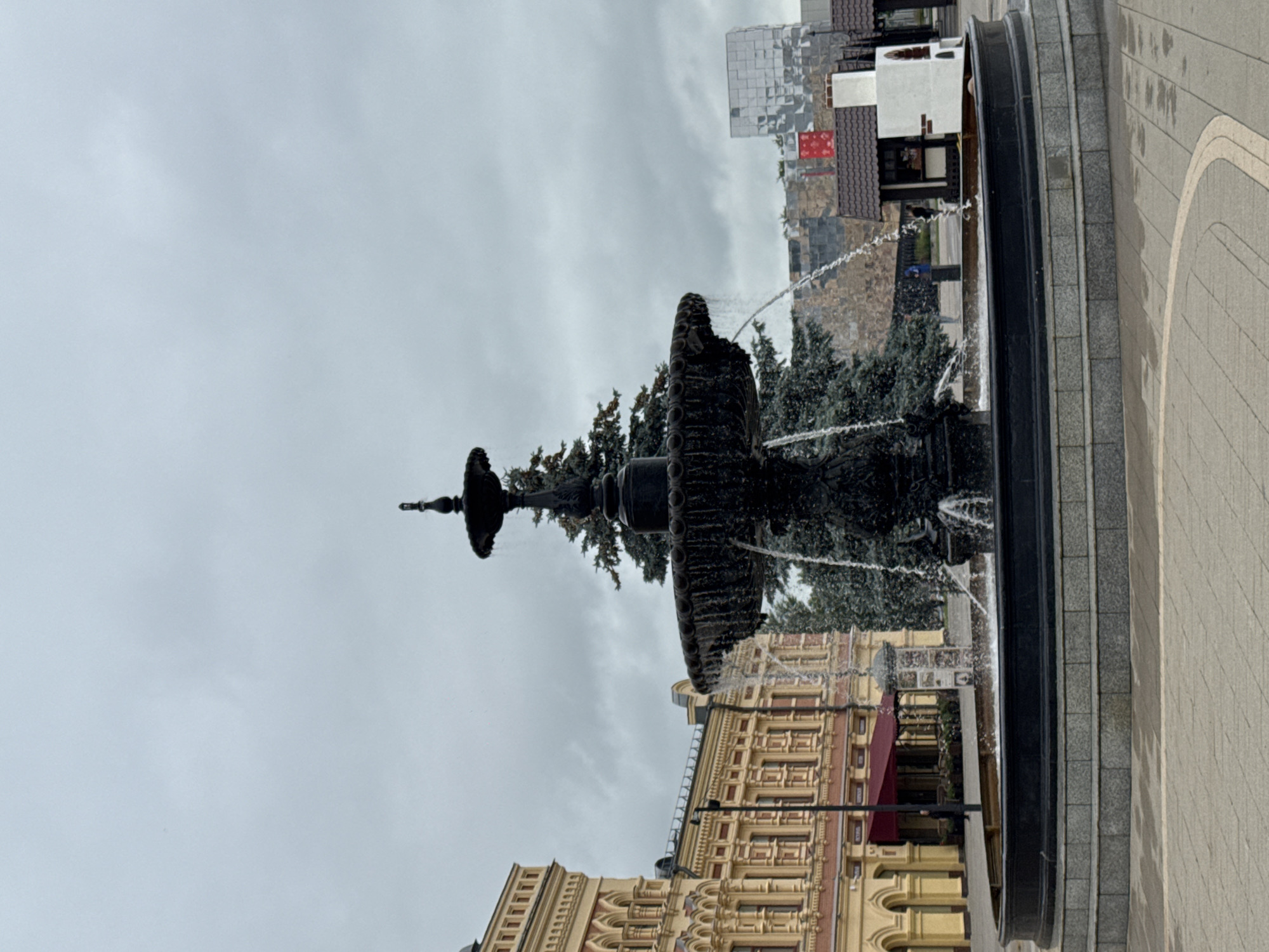 The image shows a large, ornate, dark-colored fountain in the foreground, with water cascading down its tiers. The fountain is situated in a paved public area with a patterned walkway. In the background, to the left, is a yellow building with arched windows and red awnings. To the right of the fountain, there are some smaller structures, including a white one with a brown roof and a red banner, and behind that, a modern building with a reflective, faceted facade. A tall evergreen tree is visible behind the fountain. The sky is overcast and gray. This scene is located in Nizhny Novgorod, Russia, specifically on Bolshaya Pokrovskaya Street. Two people are visible in the background near the yellow building, and one person is visible near the small structures on the right. The overall atmosphere is subdued, possibly due to the cloudy weather.