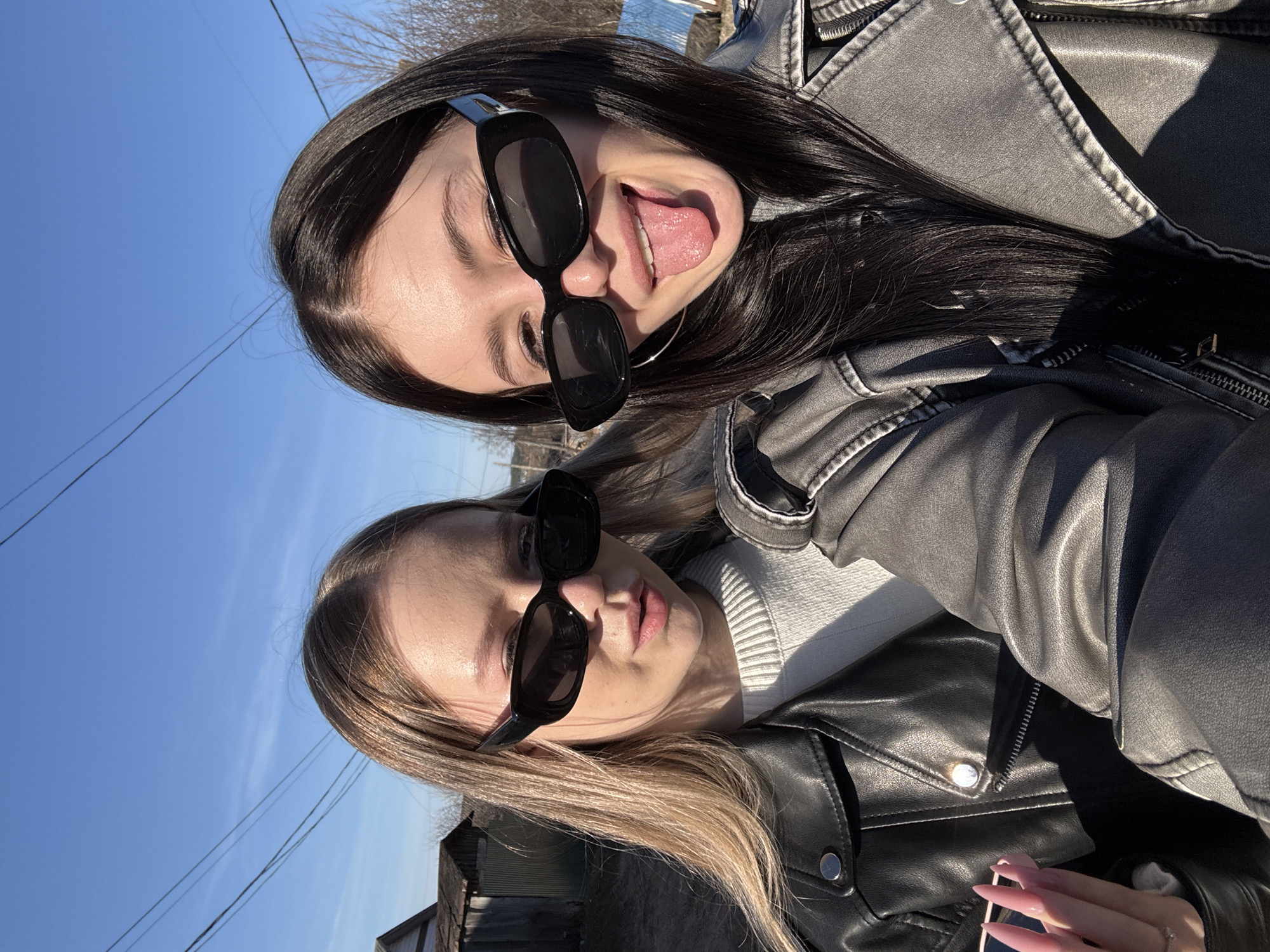 Two young women are featured in a bright outdoor selfie, captured sideways against a clear blue sky, likely in or near Фоки, Russia.
The woman closer to the bottom of the frame has long, light brown hair and is wearing a black leather jacket over a white ribbed sweater. She sports large black rectangular sunglasses and looks towards the camera with a subtle expression. Her left hand, with long, light pink or nude painted fingernails, holds a phone, partially visible at the bottom right.
Positioned above and slightly behind her, the second woman has long dark hair and is wearing a grey leather jacket that appears to have a slightly distressed or worn finish. She also wears similar black rectangular sunglasses and is playfully sticking her tongue out towards the camera. A silver hoop earring is visible on her left ear.
The background consists of a vibrant blue sky, indicative of a clear, sunny day. Thin power lines traverse the sky, and in the distance, a blurry outline of what might be a body of water or distant land is visible, along with some simple structures. The bright sunlight creates clear contrasts and highlights the textures of their clothing and hair. No visible text is present in the image. The overall atmosphere is casual and cheerful.