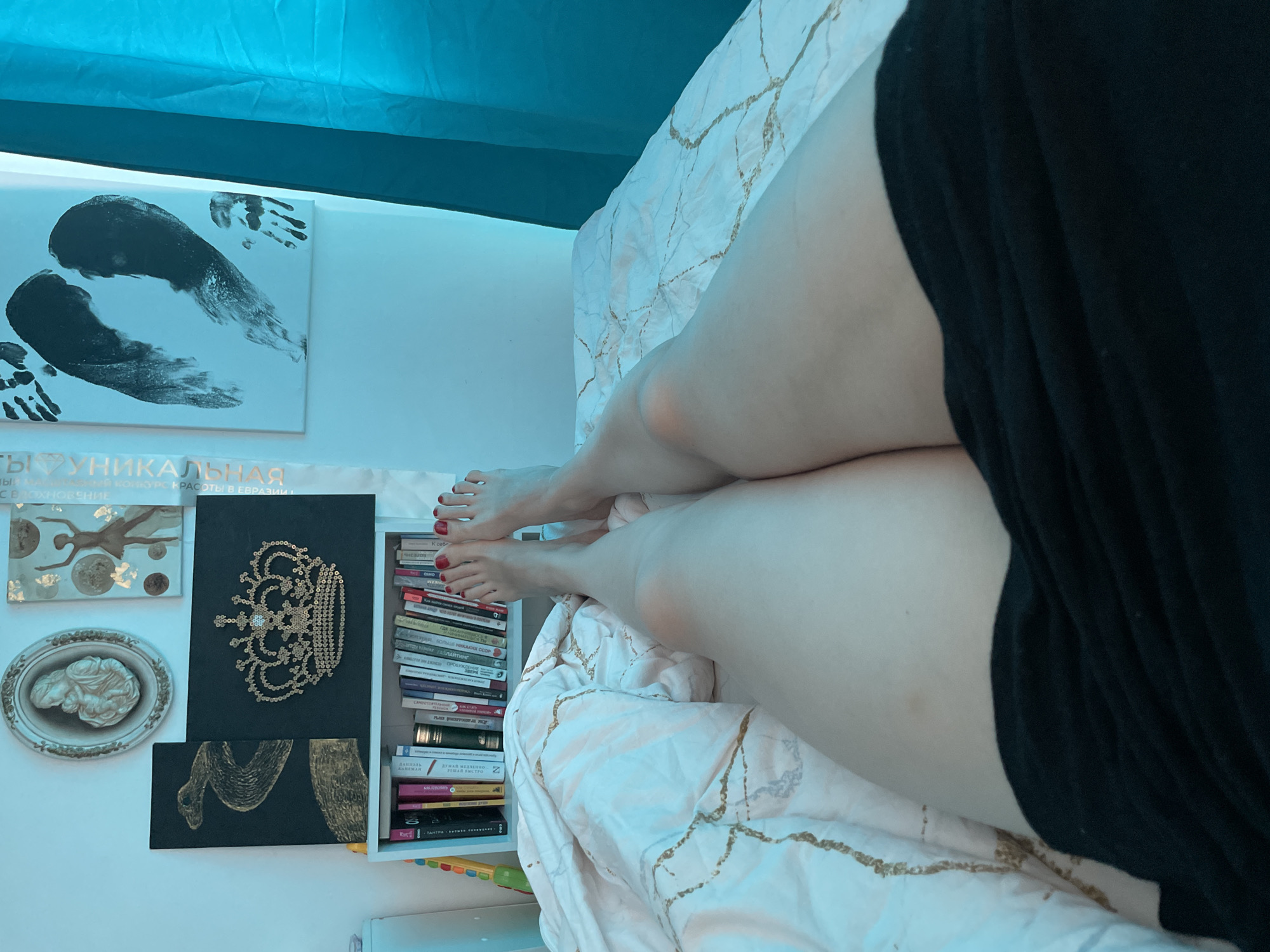 The image depicts a first-person perspective from an individual lying on a bed in a room in the city of Unknown, Unknown. The subject's bare feet, with toenails painted bright red, are positioned in the foreground, extending towards a white bookshelf filled with numerous books of varying sizes and colors. A colorful, segmented toy or object is visible on the far left of the bookshelf. Above the bookshelf, on a pale wall illuminated by a strong, cool blue light, are several decorative items. These include an oval-framed, off-white bas-relief sculpture depicting a Madonna and child (top left), a rectangular canvas with a golden ballet dancer figure (top right, with partially visible Cyrillic text), a black rectangular canvas featuring a golden coiled snake design (bottom left, above the shelf), and another black rectangular canvas adorned with a large, intricate golden crown design made of sequins or similar reflective elements (bottom right, above the shelf). To the far right, on the wall, is a white rectangular canvas displaying two large black stylized footprints or inkblots and a smaller black handprint. Vertical Cyrillic text is also visible on the wall between the crown artwork and the footprint artwork. The bed is covered with a white duvet featuring a repeating pattern of irregular gold-colored lines. The subject's thighs are visible, extending from under a black fabric garment, with a small, faint discoloration present on the subject's right thigh. A blue fabric, likely a curtain, is visible on the far right of the scene, matching the dominant cool blue ambient lighting.