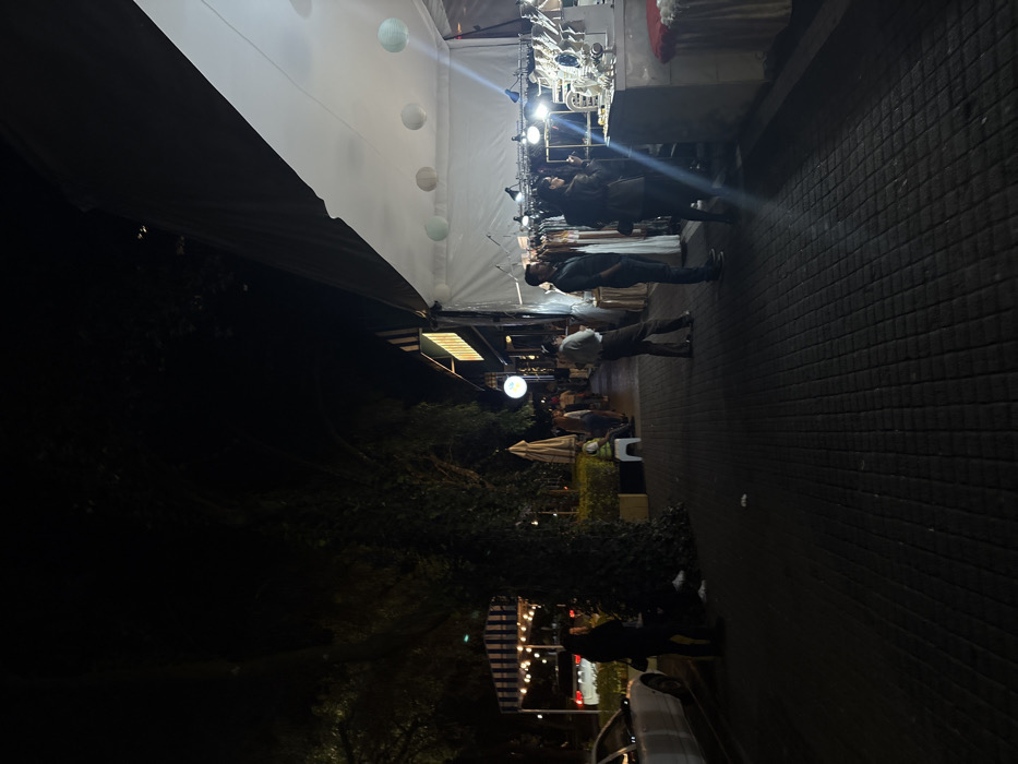 An outdoor event unfolds at night. A white tent with decorative lanterns stretches above a bustling market area. People stroll past tables, browsing the merchandise. A sidewalk with a brick pattern leads into the darker foreground. Below, trees and streetlights add to the ambiance. A car and pedestrians are visible in the lower part of the image.