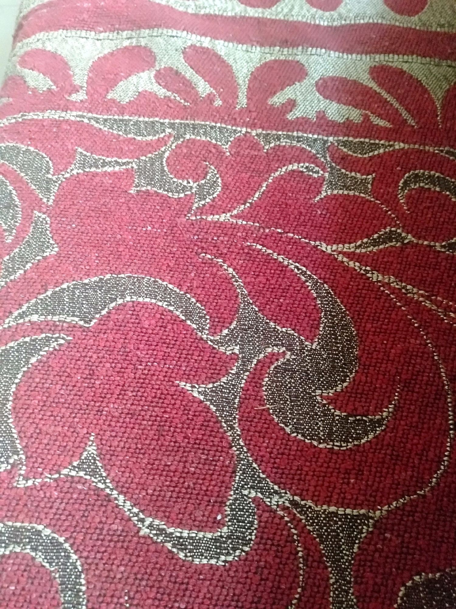 The image shows a close-up of a textured fabric with a repeating damask pattern. The fabric is primarily a deep red color, with a contrasting pattern in a silver or light grey material. The pattern consists of swirling, organic shapes that are outlined in a dark grey or black thread, giving it a three-dimensional appearance. The texture of the fabric is rough and woven, with visible threads creating a slightly raised surface. The lighting appears to be natural and diffuse, casting no harsh shadows. There are no people or distinct objects visible in the image, only the fabric itself. The specific location in Shirpur, India, cannot be determined from the visual information provided.