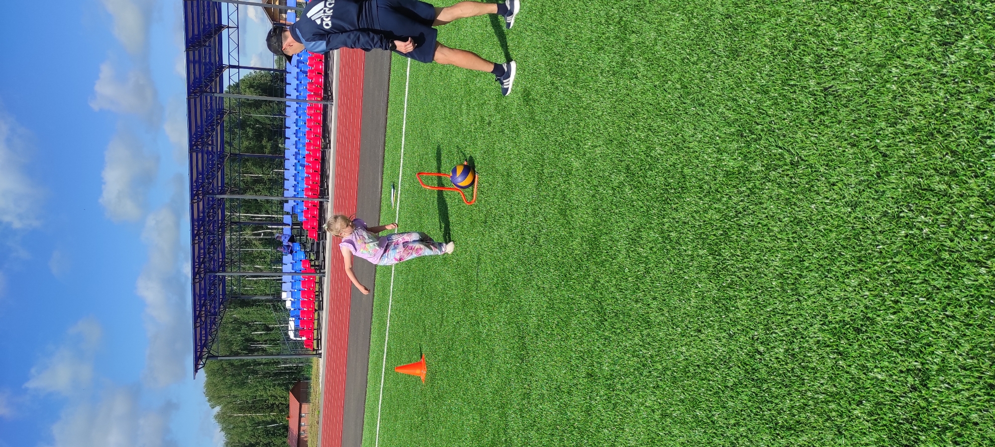 The image depicts an outdoor sports training session on a bright, sunny day in Noviy, Russia. The scene is set on a green artificial turf field, likely a soccer or athletic field, with a track running alongside. In the background, a stadium with tiered blue and red seating is visible under a clear blue sky with scattered clouds. Trees surround the field, indicating a park or sports complex setting.
The primary subjects are two people. A young girl, wearing a colorful, patterned outfit, is on the ground, appearing to be in motion, possibly during an agility drill. A man, dressed in athletic wear, including a dark blue jacket with white stripes and a cap, stands nearby, observing the girl. He is positioned further up the field.
Objects present include a volleyball resting on an orange hoop or hurdle, placed on the turf. There is also an orange traffic cone positioned on the field, suggesting an obstacle course or training setup.
The activity appears to be a training session, likely for young athletes, with the man acting as a coach or instructor. The overall mood seems positive and energetic, consistent with an outdoor sports activity. The lighting suggests it is daytime, with the sun casting shadows. There is no visible text in the image.