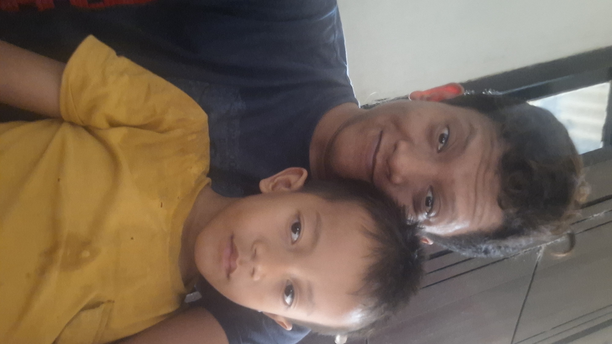 A selfie taken from a low angle shows an adult and a child from the chest up, with the adult positioned behind and to the left of the child. The adult has dark, curly hair styled upwards and is looking towards the camera with a slight smile. They are wearing a dark blue t-shirt. The child, facing the camera, has dark hair and is wearing a bright yellow t-shirt. The background appears to be indoors with a window or opening visible to the upper left, suggesting a room in a building. The overall scene is a casual, personal moment captured by the adult.