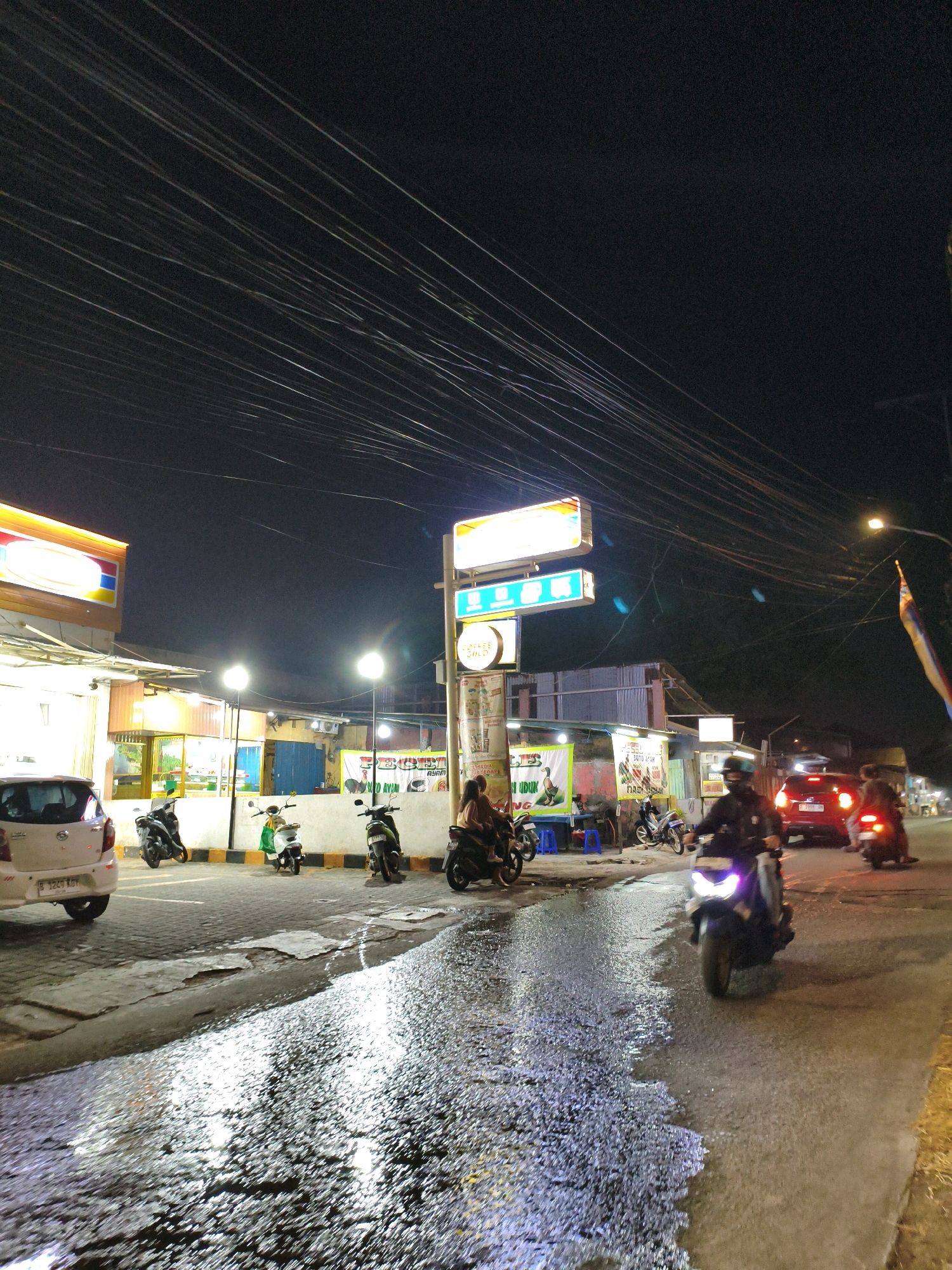 The image depicts a street scene at night in Bekasi, Indonesia, likely in an urban or suburban area. The street is wet, reflecting the lights from various establishments and street lamps. Several motorcycles are present, with some being ridden and others parked. A white car is parked on the left side of the image.
On the left, there is a brightly lit storefront, possibly a convenience store, with a prominent yellow and red sign. In front of this store, several motorcycles are parked. Further down the street, there are other shops or food stalls with illuminated signs and posters. One poster shows an illustration of a duck, and another displays text in Indonesian, possibly advertising food items.
In the foreground and mid-ground, motorcycles are moving along the street. One motorcycle in the mid-right is illuminated by its headlight and appears to be in motion. Behind it, a red car is also visible, its taillights illuminated.
The sky is dark and filled with a dense network of overhead power lines, which are a prominent feature of the upper part of the image. The overall ambiance is that of a bustling commercial street at night, possibly after rain due to the wet pavement. There are a few individuals visible, primarily on or near the motorcycles, engaged in travel or stopping at the establishments. The scene suggests a typical night in an Indonesian city where motorbikes are a common mode of transport.