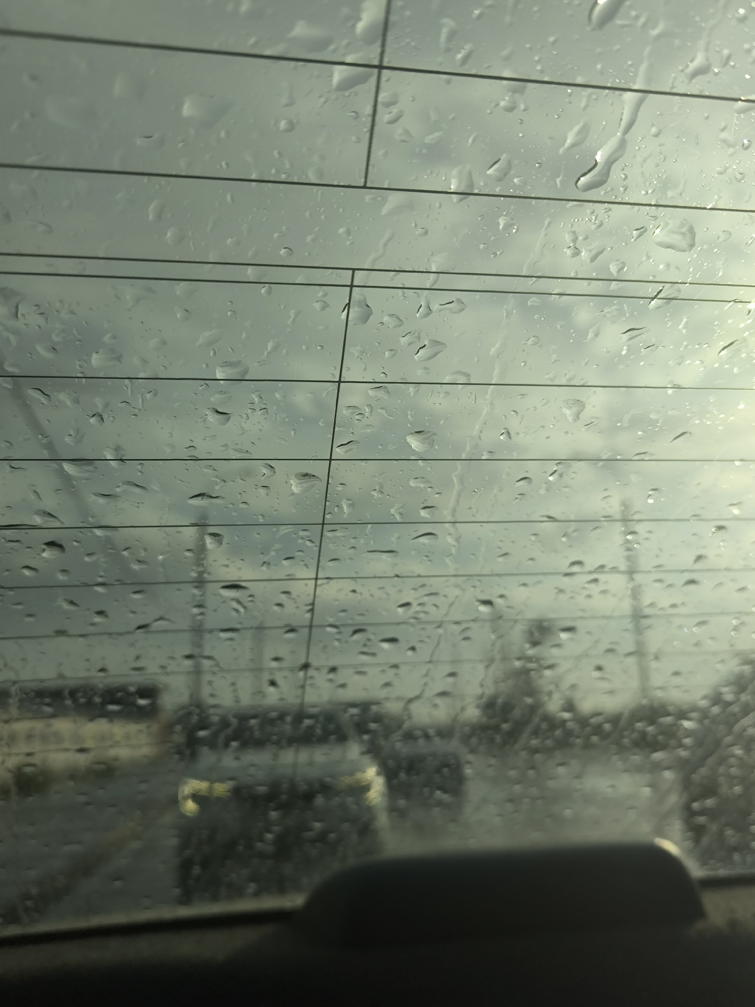 The image captures a view through a rain-streaked car window, likely the rear windshield, on a cloudy day. Horizontal defroster lines are visible across the glass, intersected by vertical lines, creating a grid-like pattern. Water droplets of varying sizes adhere to the glass, distorting the view of the outside environment.
In the background, indistinct shapes suggest vehicles and possibly buildings. The scene appears to be an urban or suburban setting, possibly on a road or in a parking lot. The overall impression is one of a wet, overcast day. The light is diffused, indicating low sunlight, possibly early morning or late afternoon.
Without specific visual cues like street signs, landmarks, or unique architectural styles, it is impossible to definitively pinpoint the location to Novosibirsk, Russia. The scene is generic and could represent a similar view in many cities experiencing rain.