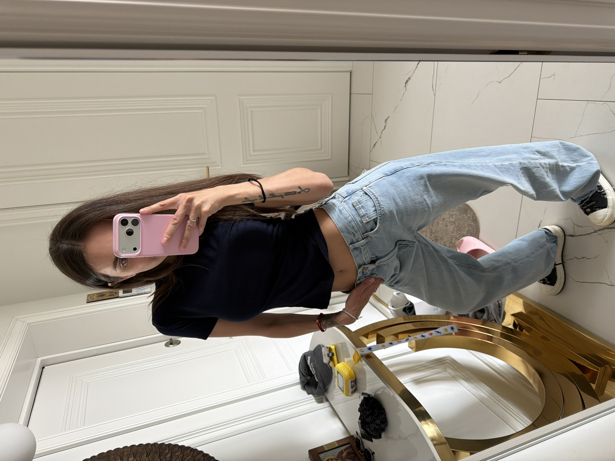 A woman is taking a selfie in a mirror. She is wearing a black crop top and light blue jeans, with black and white sneakers. Her phone is in a pink case. She is standing in what appears to be a bathroom or hallway. A white door is behind her to the left and straight ahead. The floor is white tile. A gold-colored curved table or vanity is in front of her. Other items, including a cap and sunglasses, sit on the table. The setting appears to be indoors. There is no indication of the city of Krasnoyarsk, Russia, in the image.