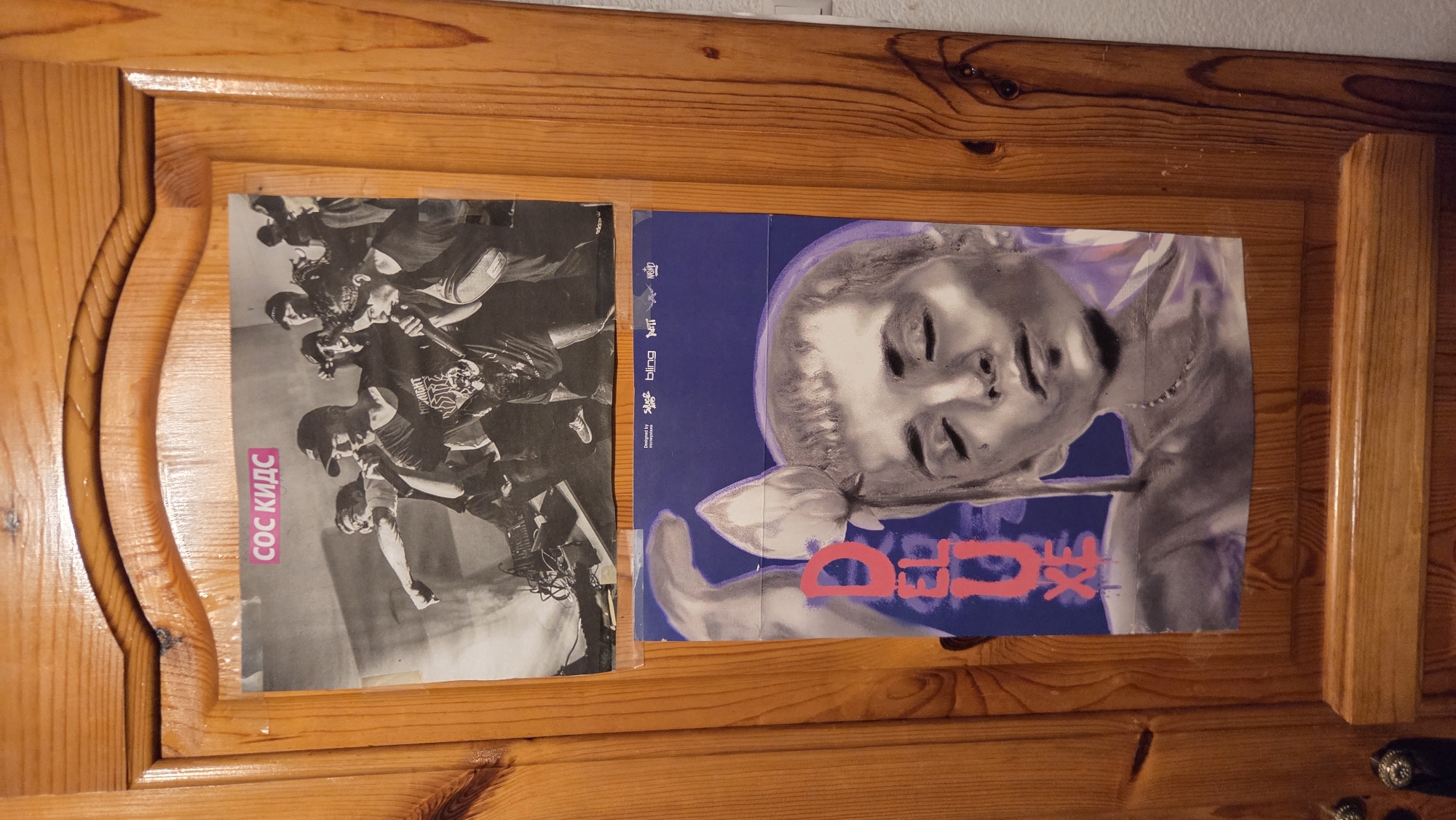 The image shows two posters on a wooden door. The top poster is a black and white photograph of a group of people holding microphones on a stage, with the text "СОС КИДС" in pink lettering at the top. The bottom poster is a portrait of a person with their eyes closed, possibly a drawing or painting, with the word "DELUXE" in large pink letters. The door is made of wood with a visible grain pattern and has some wear and tear. The scene appears to be indoors, possibly in a residential setting, given the appearance of the door.