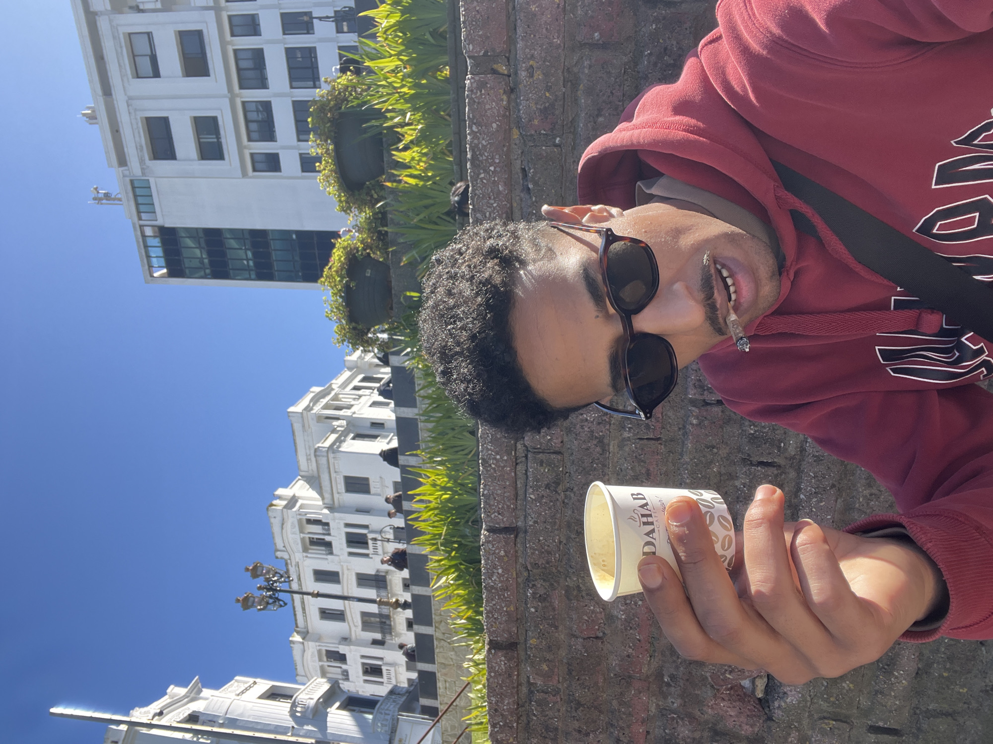 The image depicts an outdoor scene in the city of Tanger, Morocco, under a clear blue sky. In the foreground, a man takes a selfie, looking directly at the camera. He has short, curly dark hair, a mustache, and is wearing dark tortoiseshell-frame sunglasses. He is dressed in a red hooded sweatshirt with white lettering on the chest and a light-colored shirt underneath. A dark strap from a bag crosses his chest. His left hand holds a white disposable paper cup, partially filled with a light-colored liquid, bearing the logo "DAHAB CAFE." A lit cigarette is positioned between his lips.
Immediately behind the man is a weathered brick wall, above which cultivated green foliage, possibly succulents or shrubs, grows in a garden bed or planters. Sunlight brightly illuminates these plants. In the background, several multi-story buildings are visible. To the left, a large, ornate white building with classical architectural elements, including balconies and dark-framed windows, stands beside a decorative black street lamp. Further right, another prominent white building with both standard rectangular windows and sections of dark, reflective glass is visible. Antennas or other structures are present on its rooftop. Several indistinct human figures are visible on a higher level or pathway between the foliage and the ornate white building.