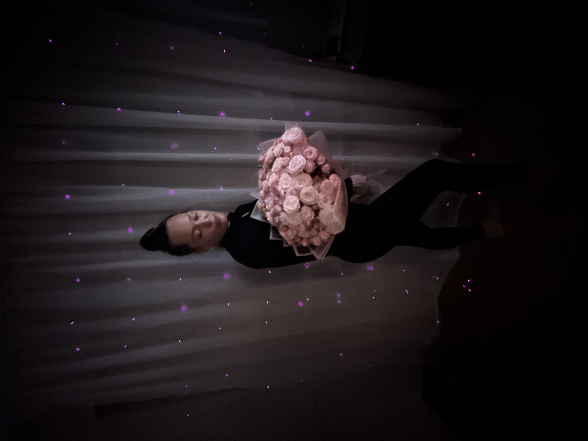 A woman lies on her back, holding a bouquet of pink roses. She is dressed in black and has her hair pulled back in a bun. The background is a dark room with what appear to be strings of lights, possibly Christmas lights. The scene is in Chaikovsky, Russia.