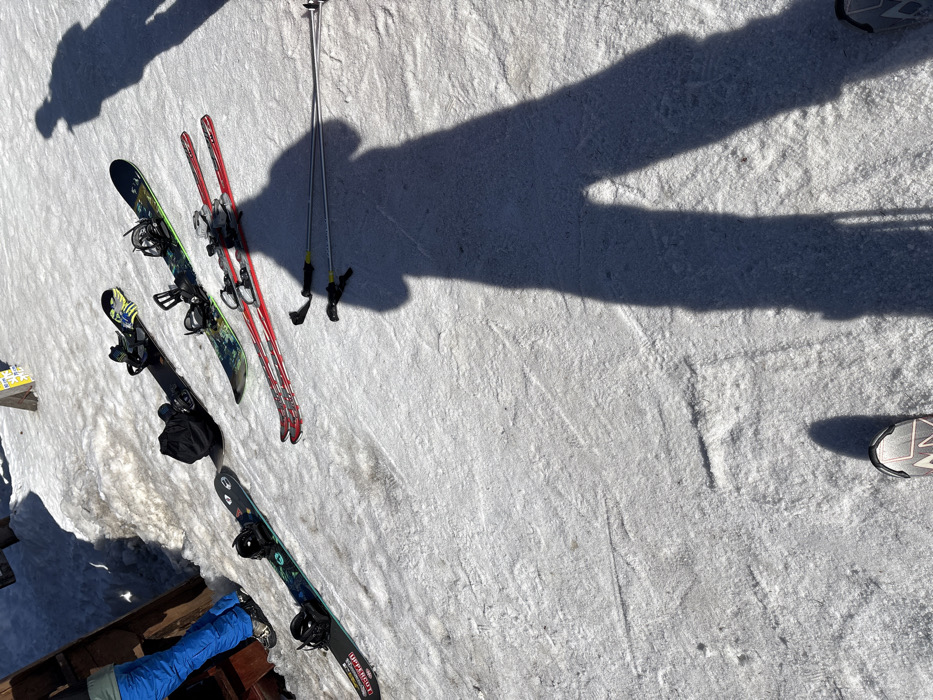 The image displays a snow-covered outdoor area, consistent with a ski slope or resort in Bistritsa, Bulgaria, under bright sunlight, evidenced by prominent shadows. Resting on the snow are three snowboards: two predominantly black, one of which has a black backpack placed on it, and a third snowboard with green and black patterns, all equipped with bindings. Adjacent to the snowboards are a pair of red skis with silver bindings and a pair of silver ski poles. To the far left, the lower legs of an individual wearing blue ski pants and dark footwear are visible, standing next to a brown wooden structure partially obscured by snow. Two distinct human shadows are cast across the snow; a large shadow belonging to the photographer occupies the foreground, with the photographer's dark and white shoes visible at the bottom edge, and a smaller shadow is present in the upper right quadrant. The snow surface exhibits various tracks and compacted patches from previous activity.