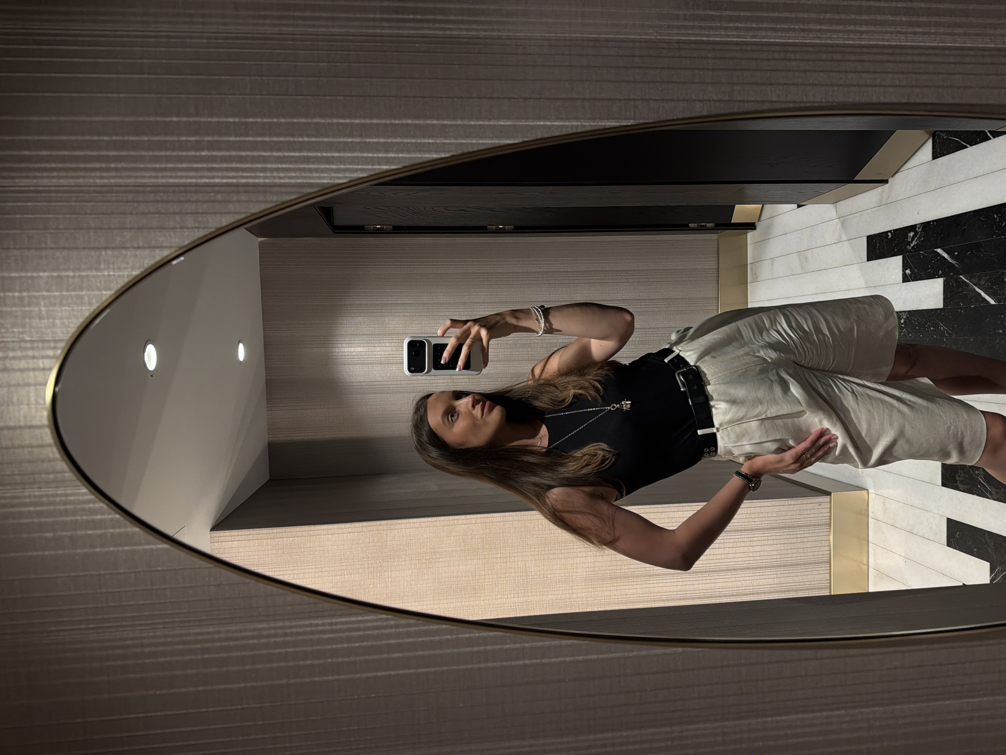 In Budva, Montenegro, a woman takes a selfie in a narrow hallway with textured wallpaper. She is wearing a black sleeveless top, a black belt, and off-white shorts. A silver necklace with a pendant hangs around her neck, and she is also wearing a watch and bracelet on her left arm. The hallway features a patterned floor with white, black, and marble tiles, and a dark wooden door is visible on the right side. The lighting is dim, with two recessed lights visible on the ceiling.
