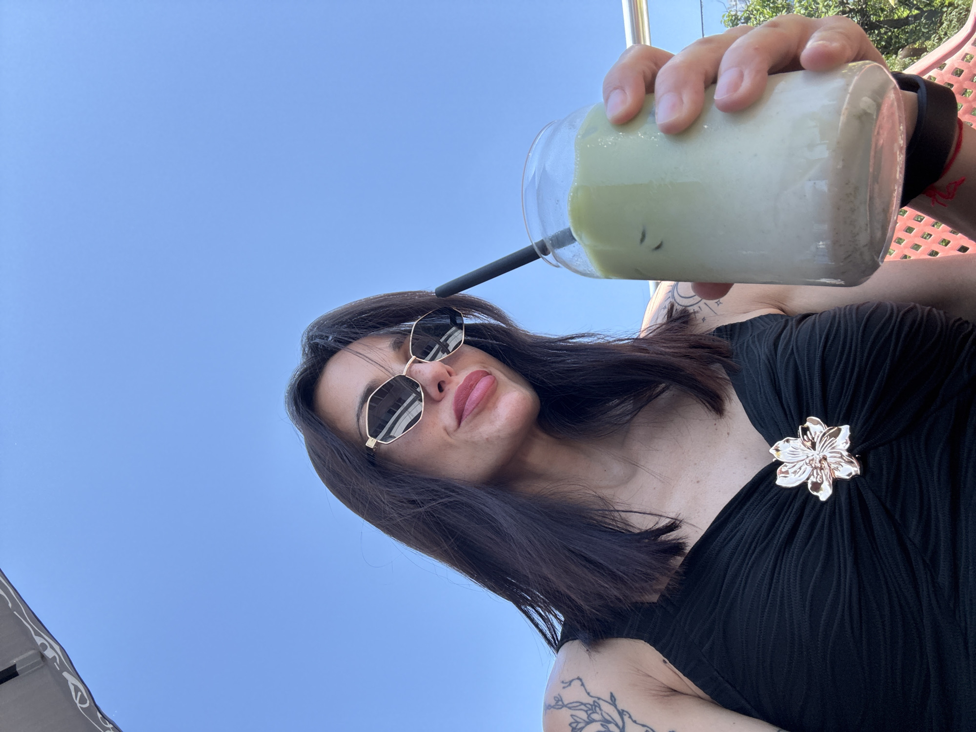 A woman is holding a glass of a light green and white beverage with a black straw against a blue sky background. She is wearing gold-framed sunglasses, a black sleeveless top with a gold flower brooch, and has dark shoulder-length hair. Tattoos are visible on her arm and chest. The beverage appears to be iced. The photo is taken from a low angle, looking up at the subject. Parts of a red metal structure are visible. The location is Pleven, Bulgaria.