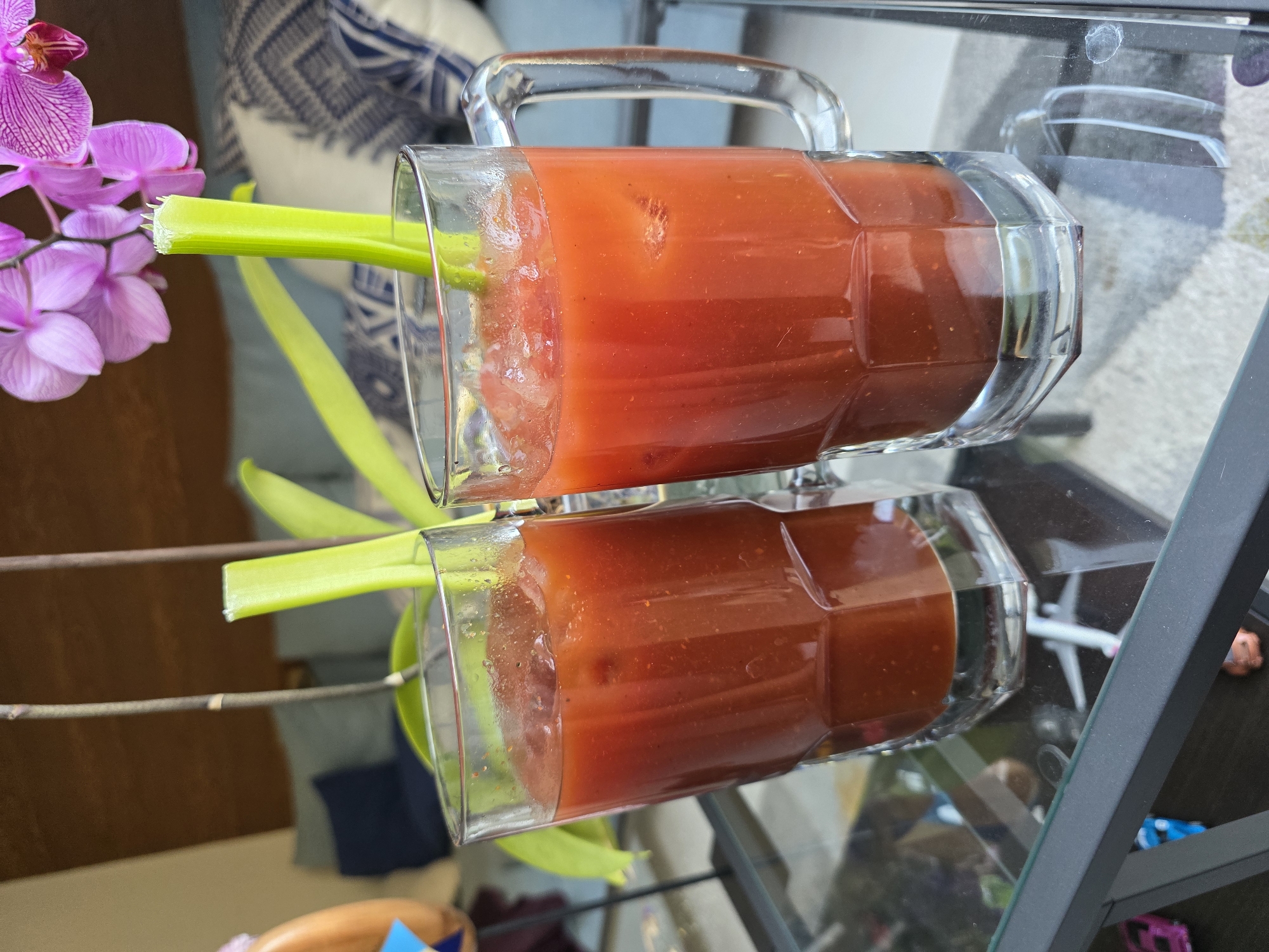 A close-up shot depicts a glass mug filled with a reddish-orange liquid, likely tomato juice or a Bloody Mary, with a celery stalk sticking out. The mug sits on a reflective surface, possibly a glass table, creating a mirror image below. To the left of the mug, partially visible, is a pink orchid. The background shows a blurred interior, suggesting a living room or dining area with a pillow in the upper-left corner. The mug has a clear handle, and the liquid appears to have some sediment or pulp. Further in the background and visible through the transparent surface are an airplane and toy cars, located inside a glass table, suggesting that the image was taken in Puebla, Mexico.