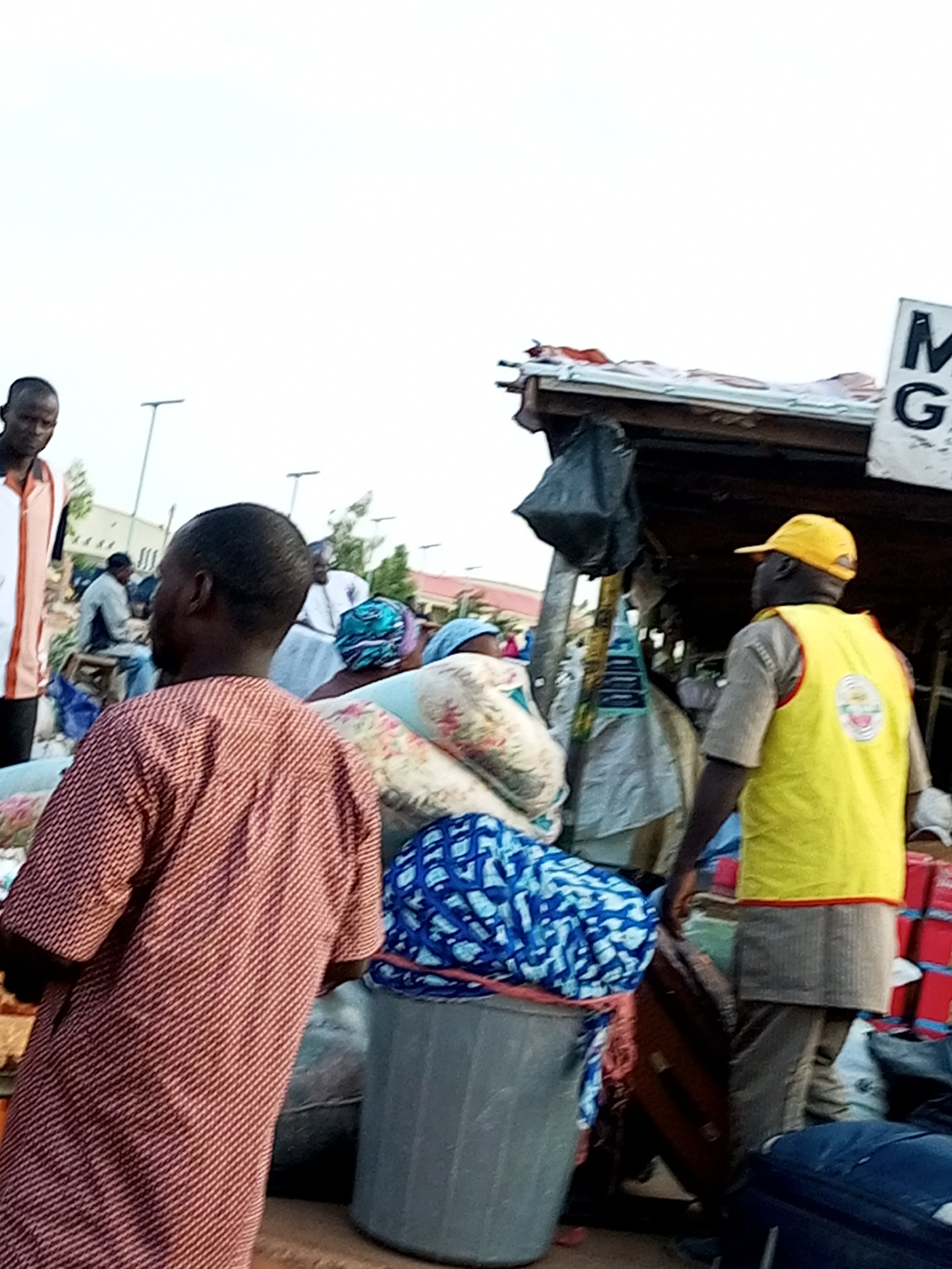 The image depicts a busy outdoor market scene, likely in Gombe, Nigeria, characterized by a crowd of people and various goods. In the foreground, a man wearing a patterned red and white traditional garment faces away from the camera, his back to the viewer. To his right, another man, wearing a yellow vest over a light brown shirt and a yellow cap, stands facing away from the camera. The yellow vest has a circular emblem on the back. Behind them, several other individuals are visible, some partially obscured, engaged in market activities or passing through.
The environment suggests a market setting with stalls and overhead structures. A black plastic bag hangs from one of the structures. Stacks of red boxes are visible to the right, and several large bags and bundles of fabric, some with vibrant blue and white patterns, are piled up. A large gray plastic bin is situated in the lower right quadrant of the image. The background reveals more market stalls, buildings, and what appear to be streetlights, indicating an urban or semi-urban location. The lighting suggests it is daytime, with bright, possibly overcast, sky. The overall impression is one of a lively and active marketplace.